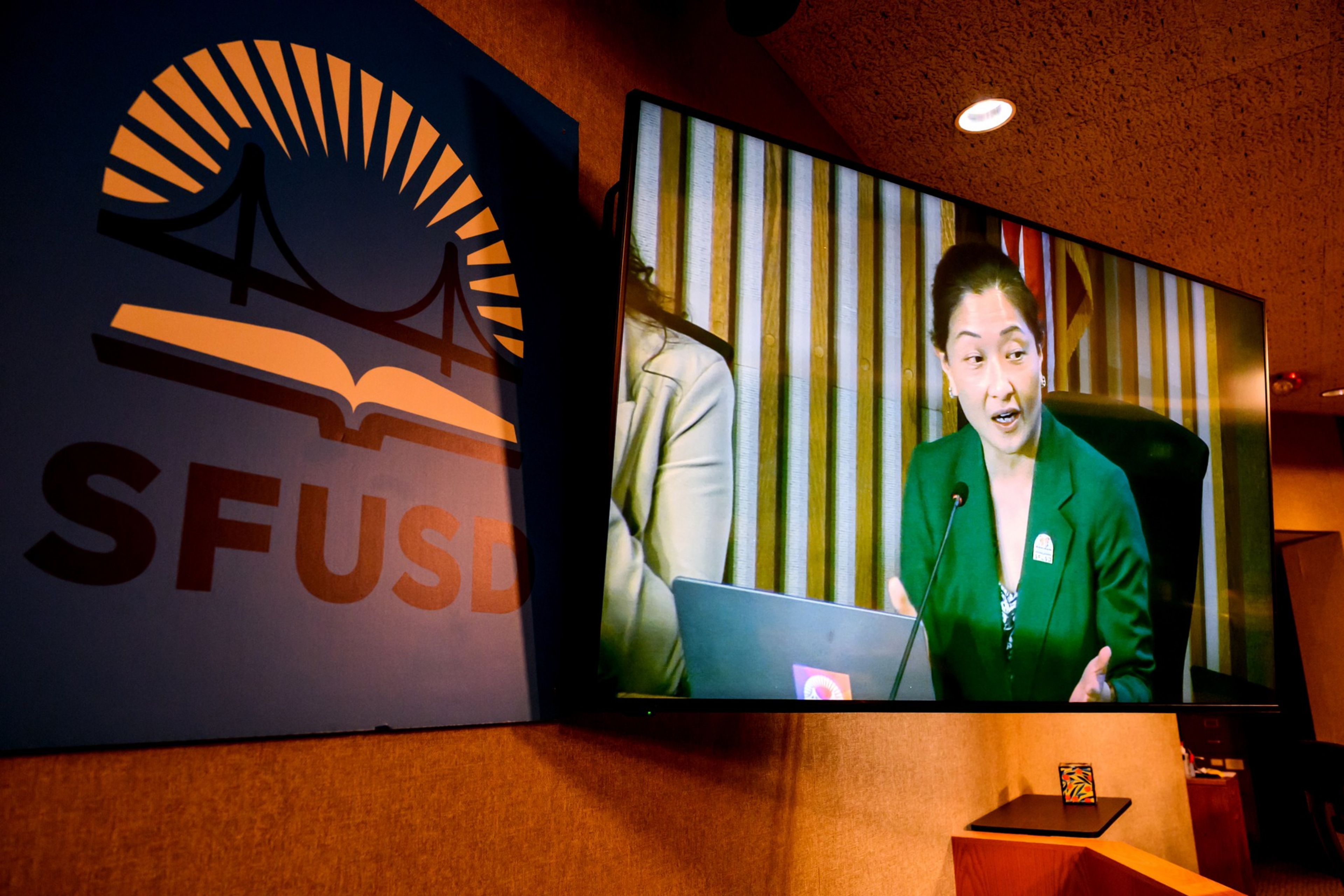 A woman in a green blazer speaks into a microphone, shown on a large screen next to a blue sign with an orange bridge and book logo labeled "SFUSD."