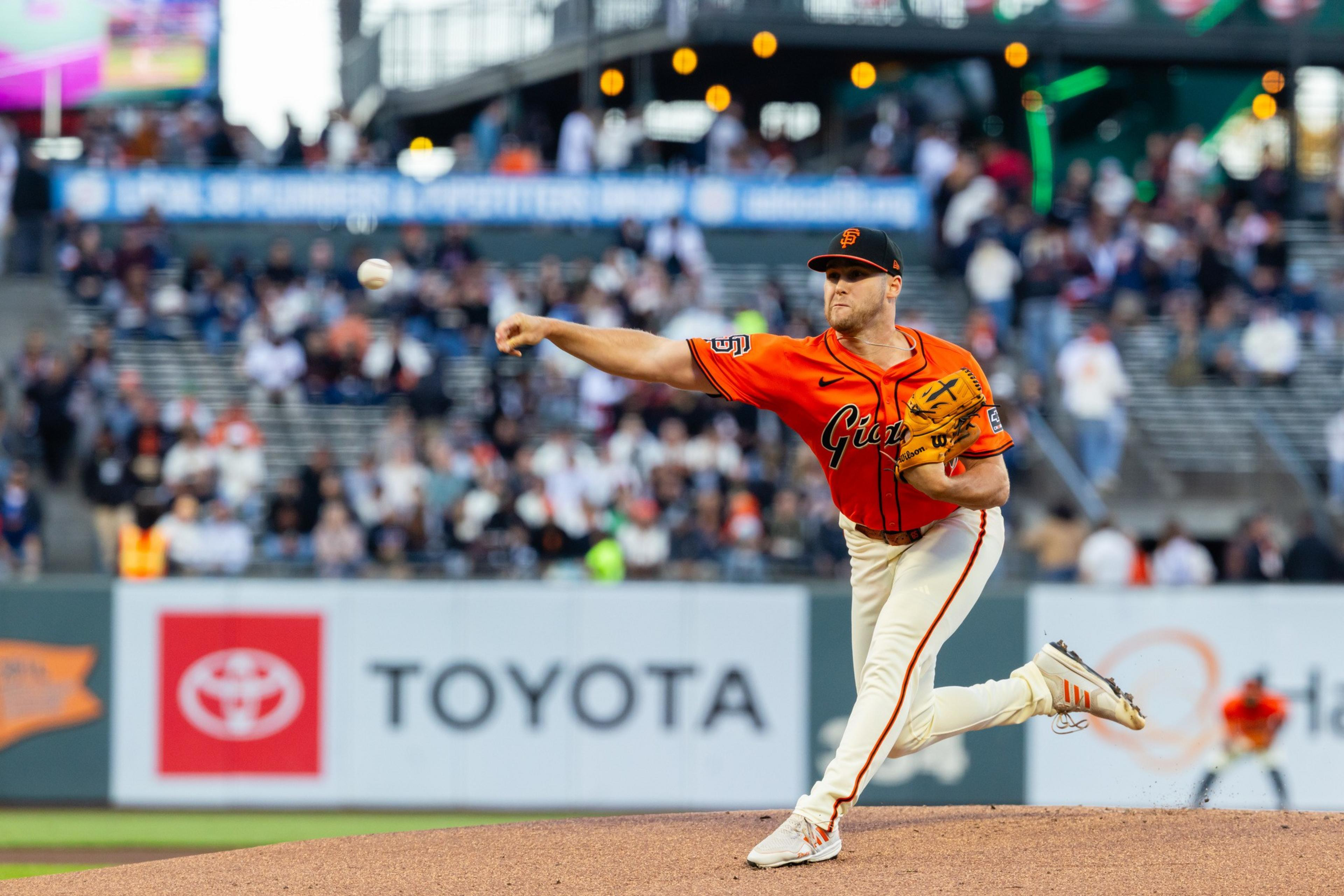 A baseball pitcher in an orange Giants uniform is mid-throw on the mound with a crowd in the blurred stadium background.