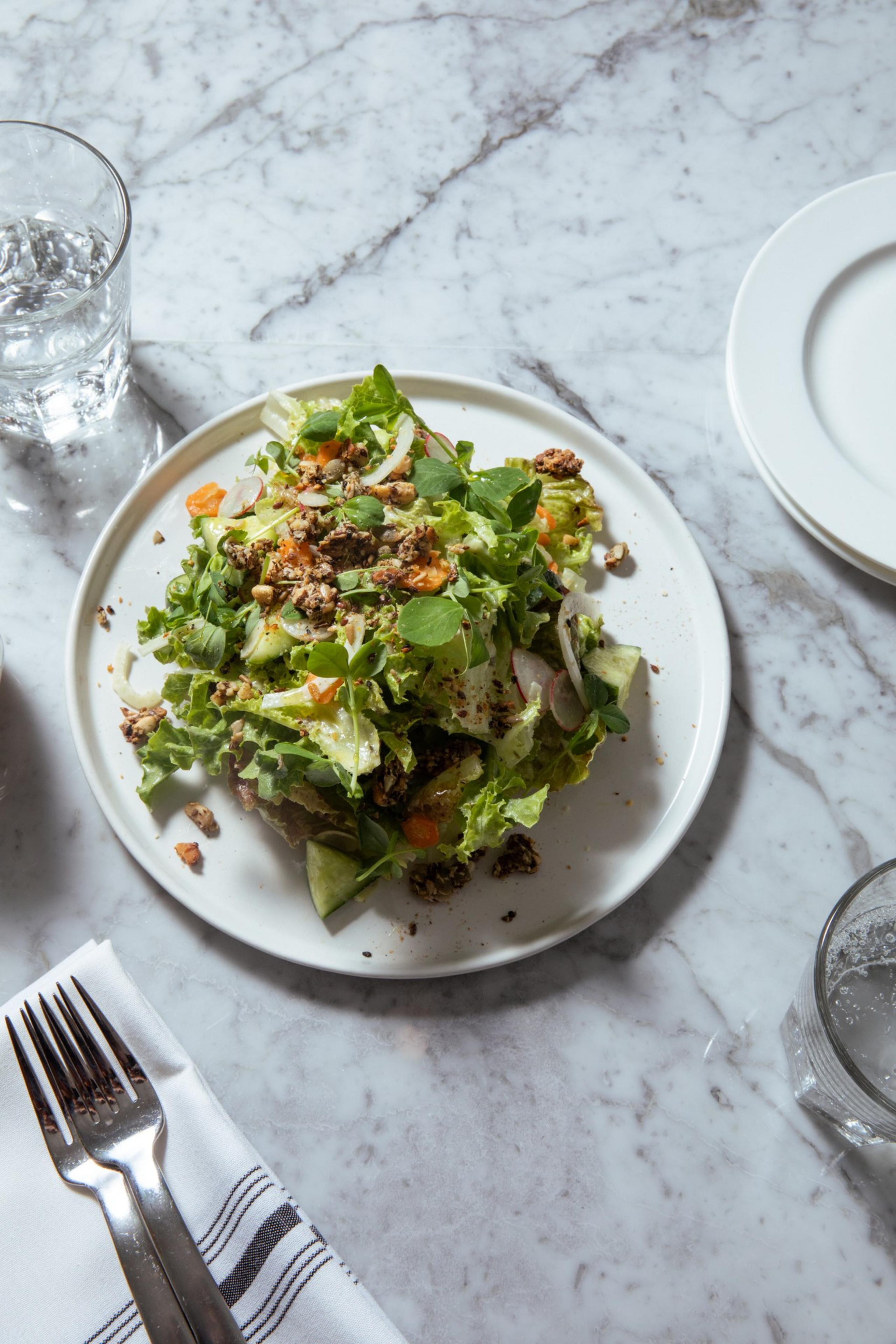 A fresh green salad with nuts and diced vegetables sits on a white plate on a marble table, accompanied by a glass of water and cutlery on a napkin.