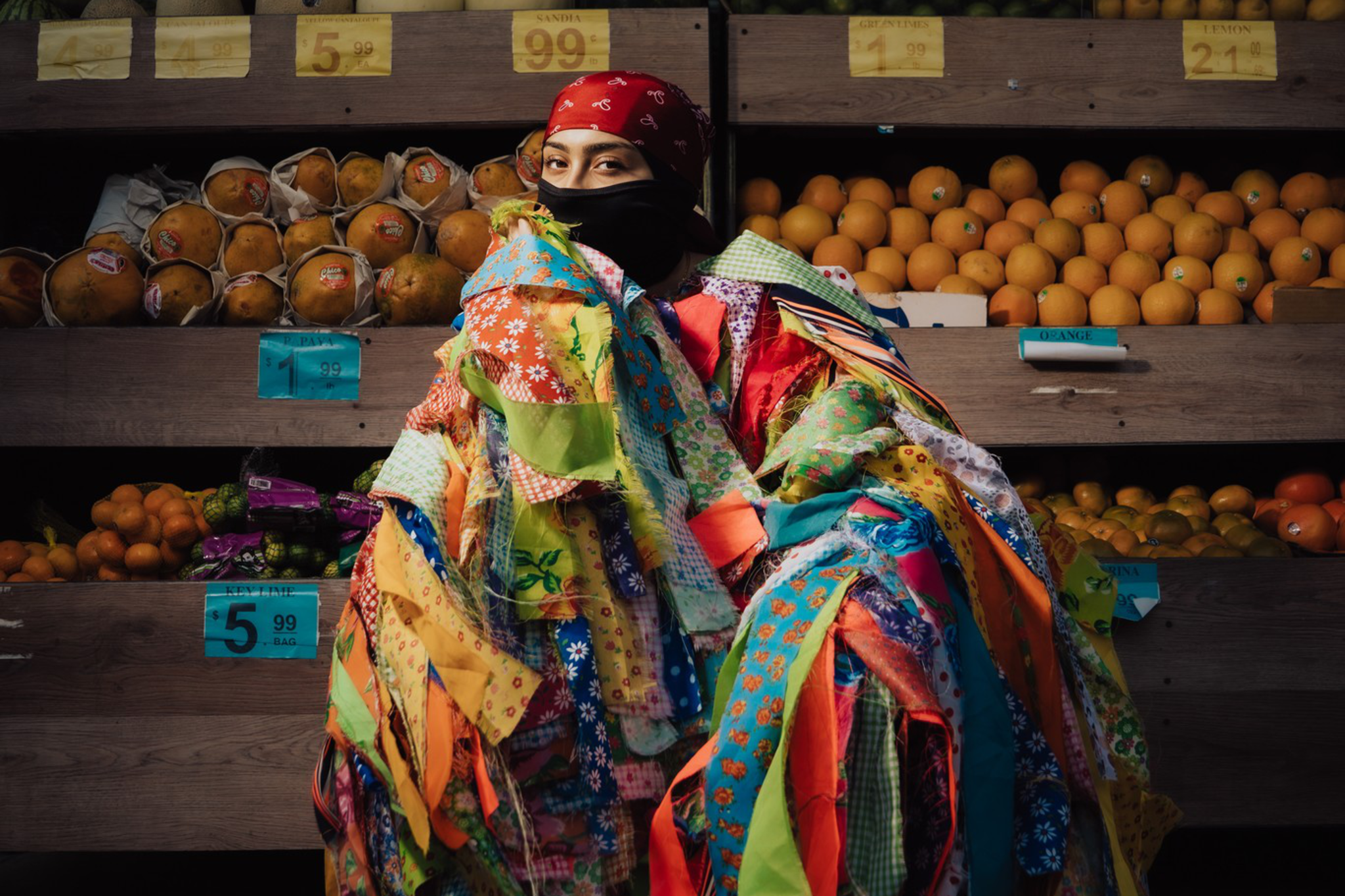 A person wrapped in a colorful patchwork quilt sits in front of fruit stands displaying cantaloupes, oranges, and other produce.
