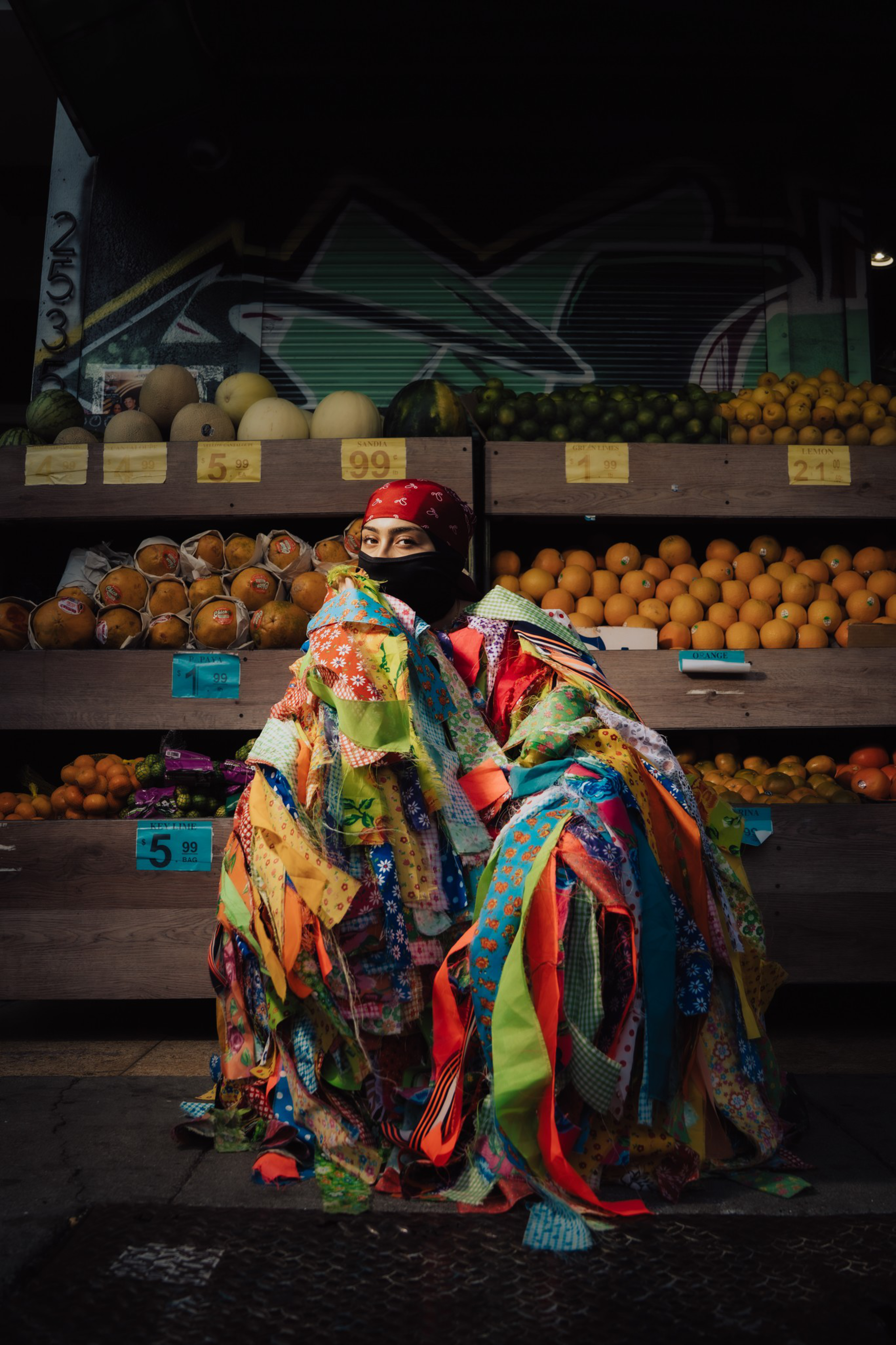 A person wrapped in a colorful patchwork quilt sits in front of fruit stands displaying cantaloupes, oranges, and other produce.