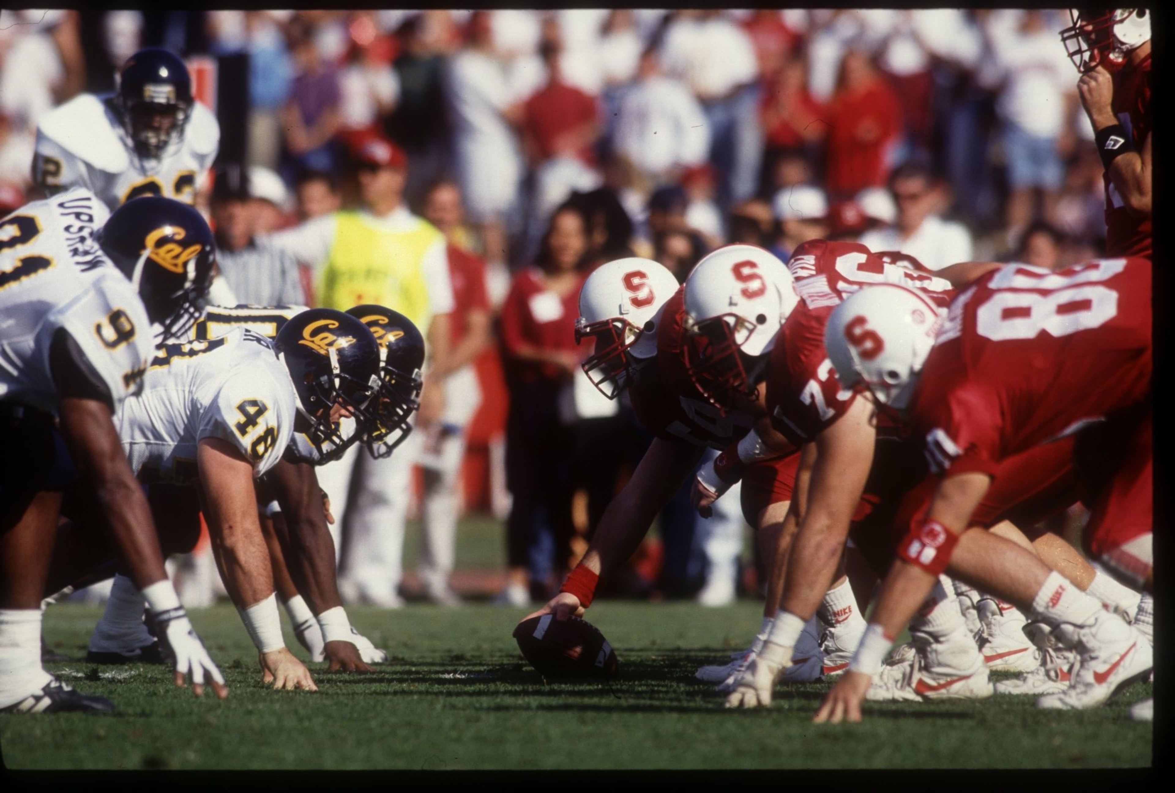 Two American football teams, one in white and navy and the other in red and white, face off on the line of scrimmage, ready for the snap.