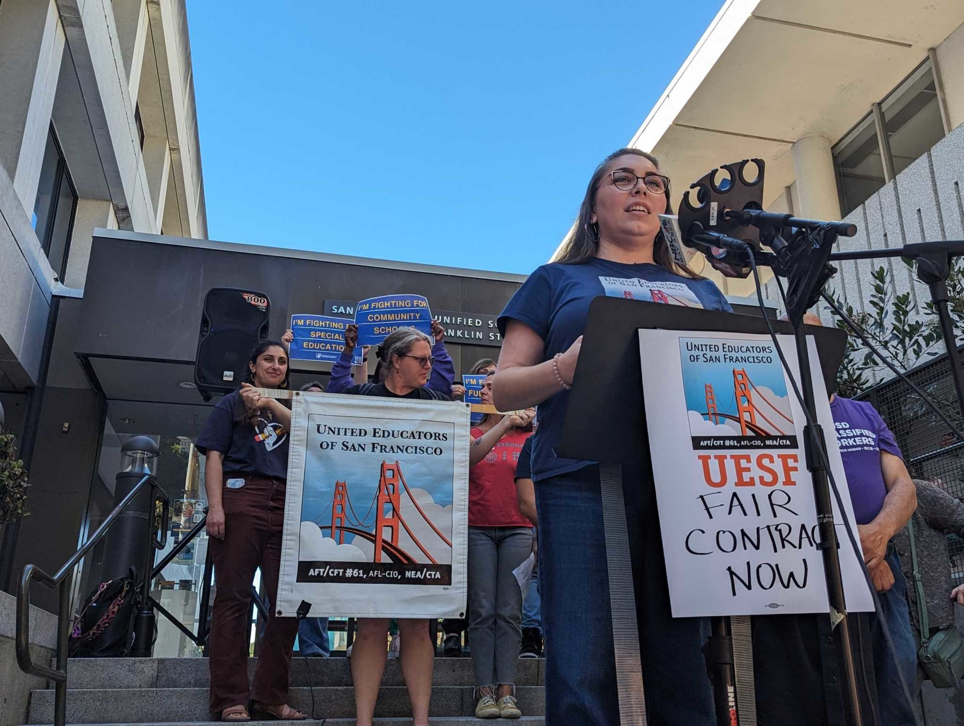 A woman speaks at a podium outdoors with a sign reading "UESF FAIR CONTRACT NOW." Several people stand behind her holding signs, in front of a modern building.