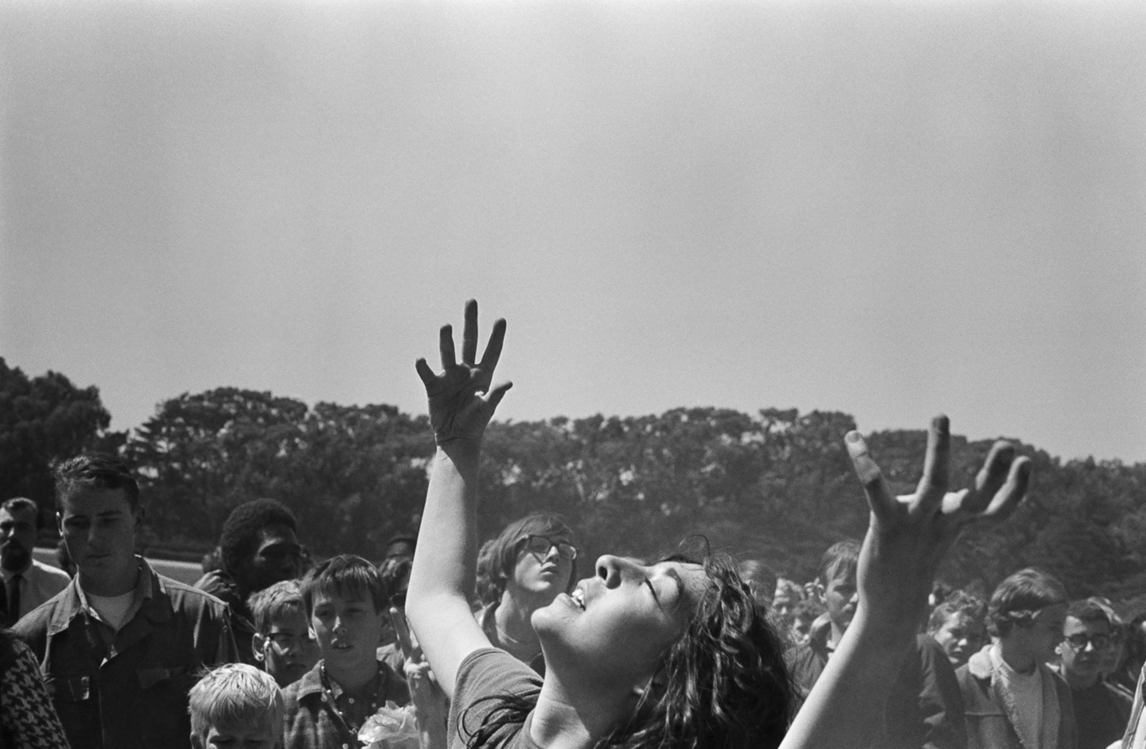A woman stands among a crowd outdoors, her face lifted skyward, eyes closed, and arms stretched up in an expressive gesture, surrounded by trees and people.
