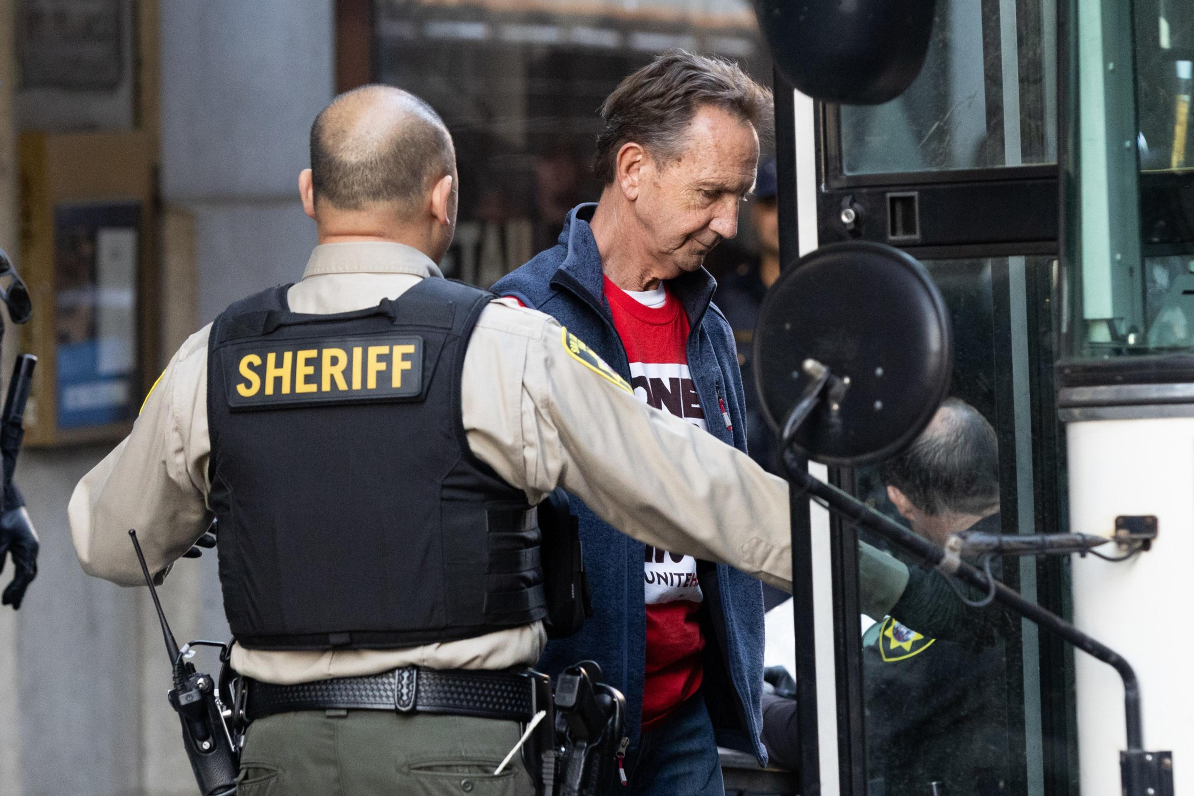 A sheriff in uniform escorts a man in a red shirt and blue jacket onto a bus. The scene appears to be in an urban setting.