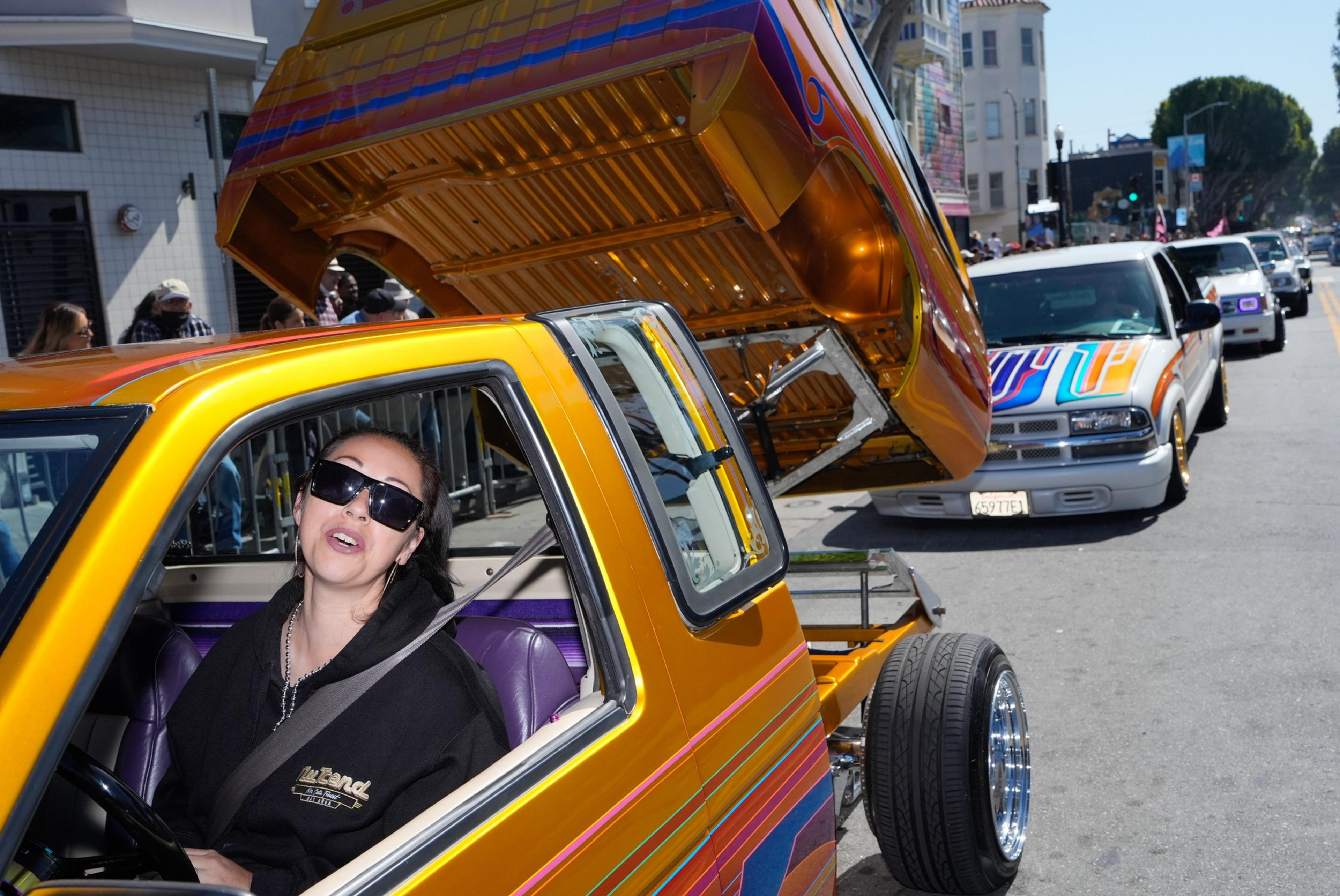 A person wearing a dark shirt and sunglasses rides in a custom pickup truck in a parade.