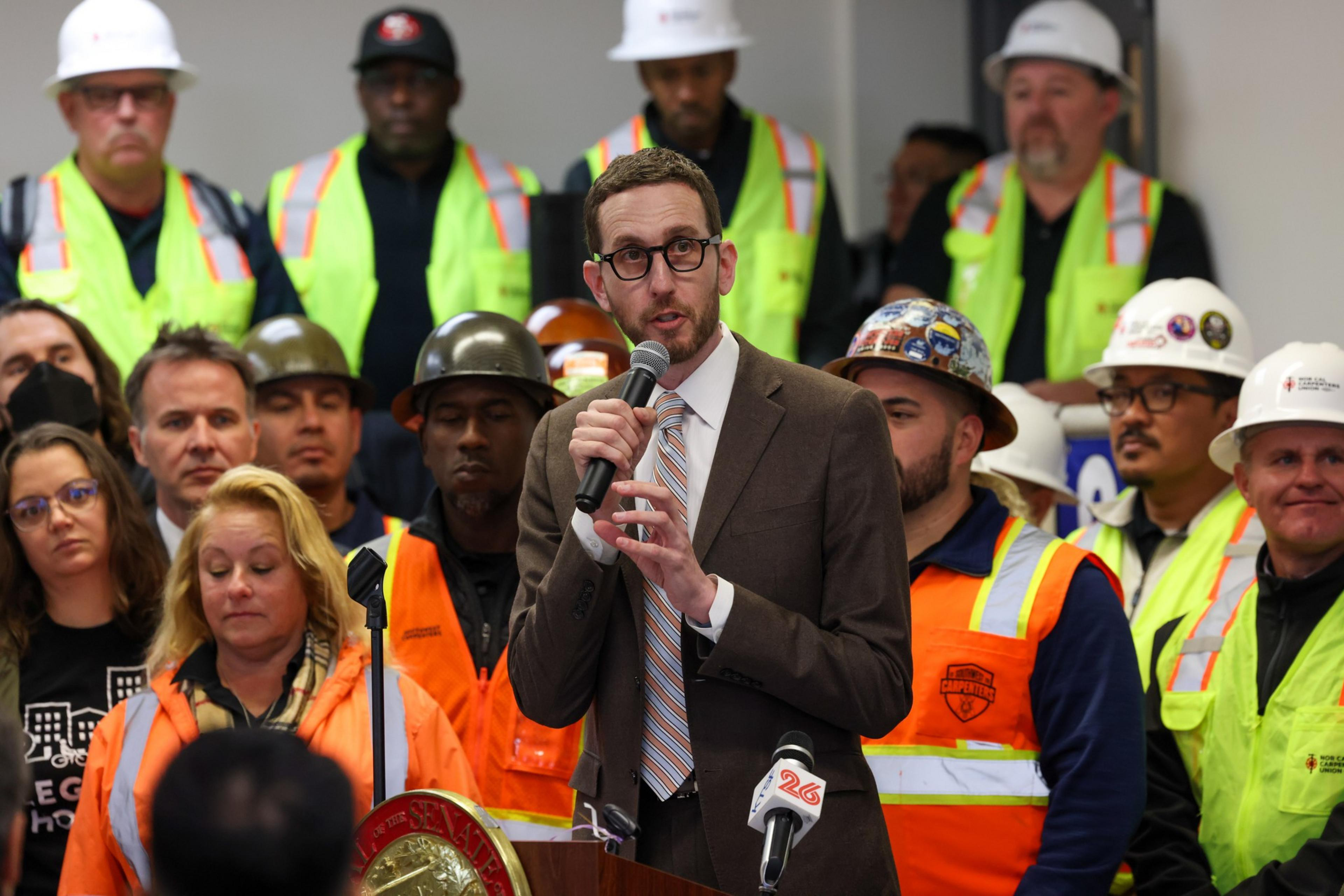 A man in a suit and tie speaks at a microphone, surrounded by construction workers wearing safety vests and helmets, inside a room.