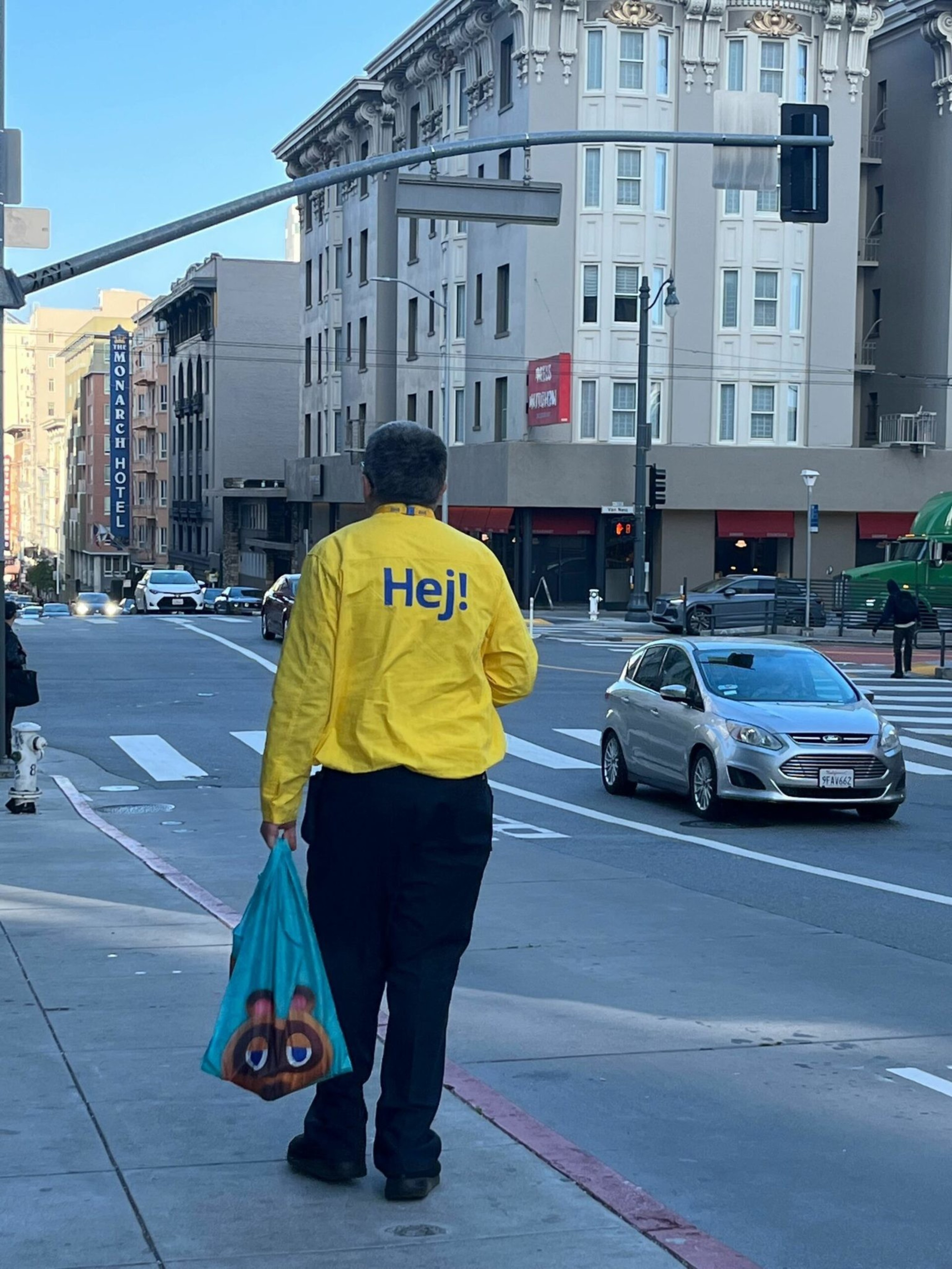 A person wearing a bright yellow jacket with "Hej!" on the back carries a blue bag with a colorful character walking along a city sidewalk.