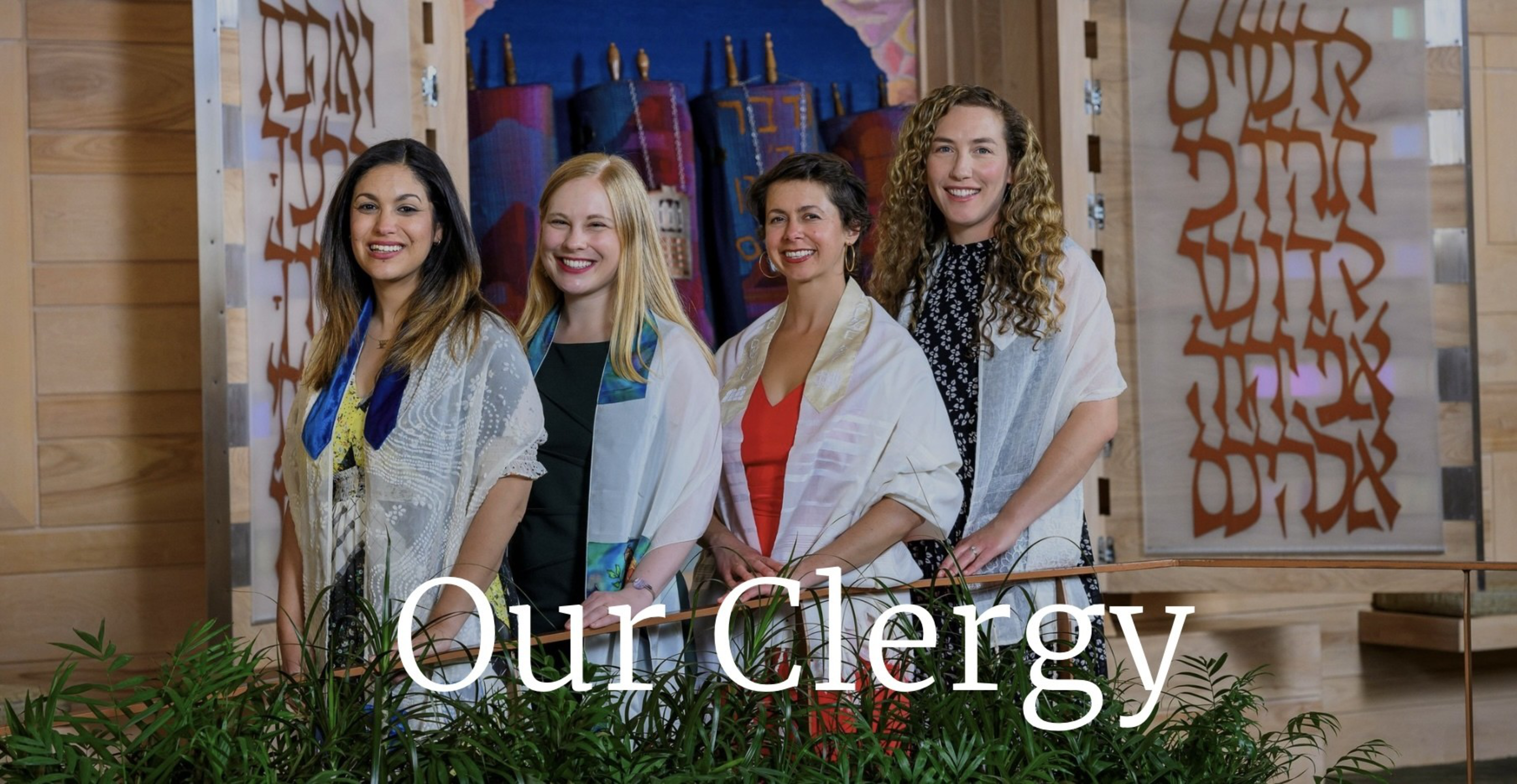 Four women stand side by side, smiling. They are wearing shawls and standing in front of a colorful backdrop with artistic designs and Hebrew letters.