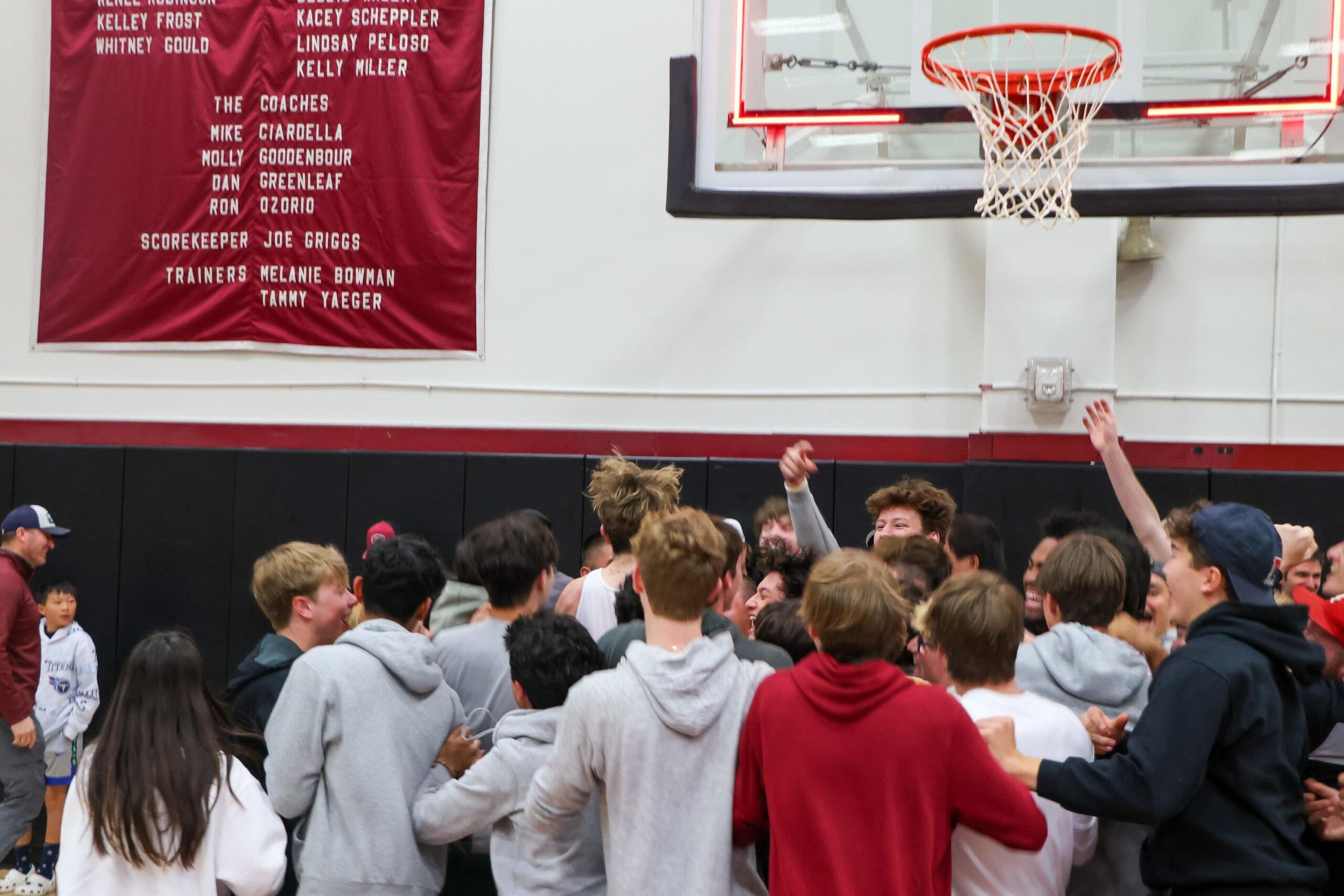 A large group of excited young people gather under a basketball hoop inside a gym, some raising their hands in celebration.