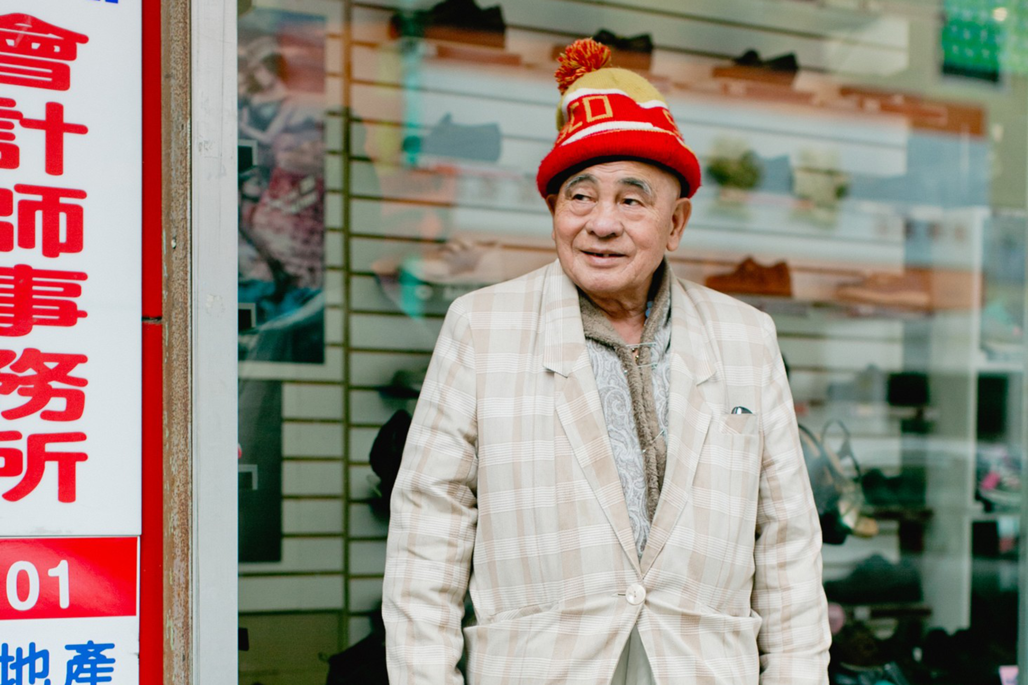 An elderly man wearing a red and yellow knitted hat, a beige plaid blazer, light trousers, and black shoes stands outside a store with shoes displayed in the window.