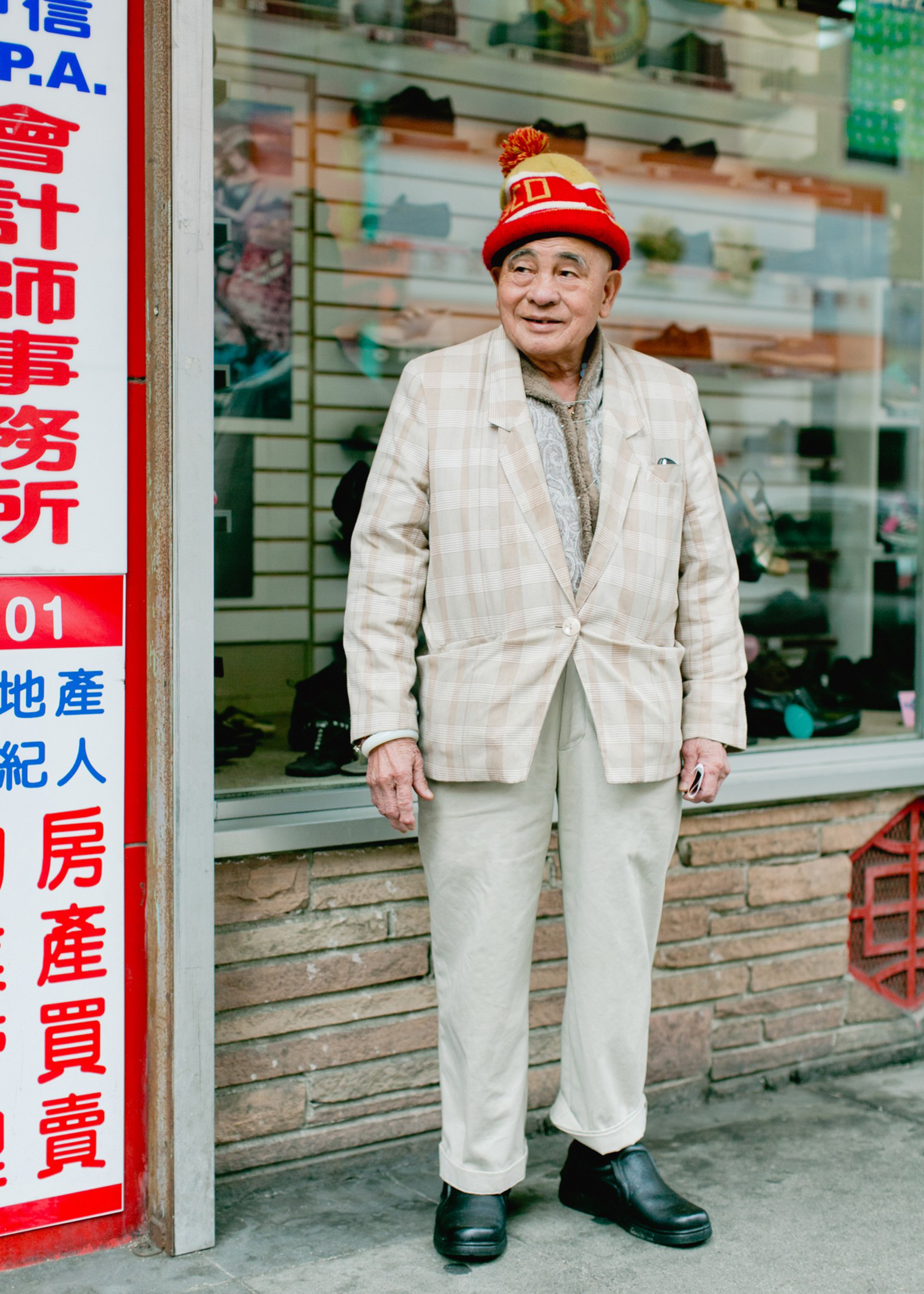 An elderly man wearing a red and yellow knitted hat, a beige plaid blazer, light trousers, and black shoes stands outside a store with shoes displayed in the window.