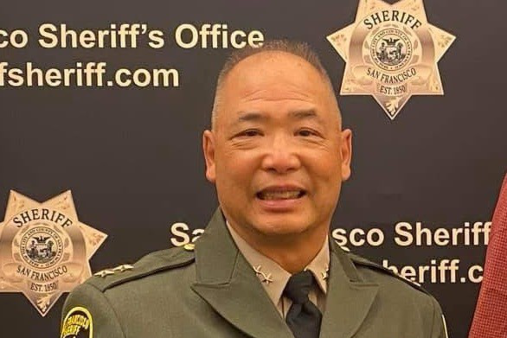 A uniformed officer smiles, standing in front of a San Francisco Sheriff's Office backdrop. The uniform is dark with badges and a name tag.