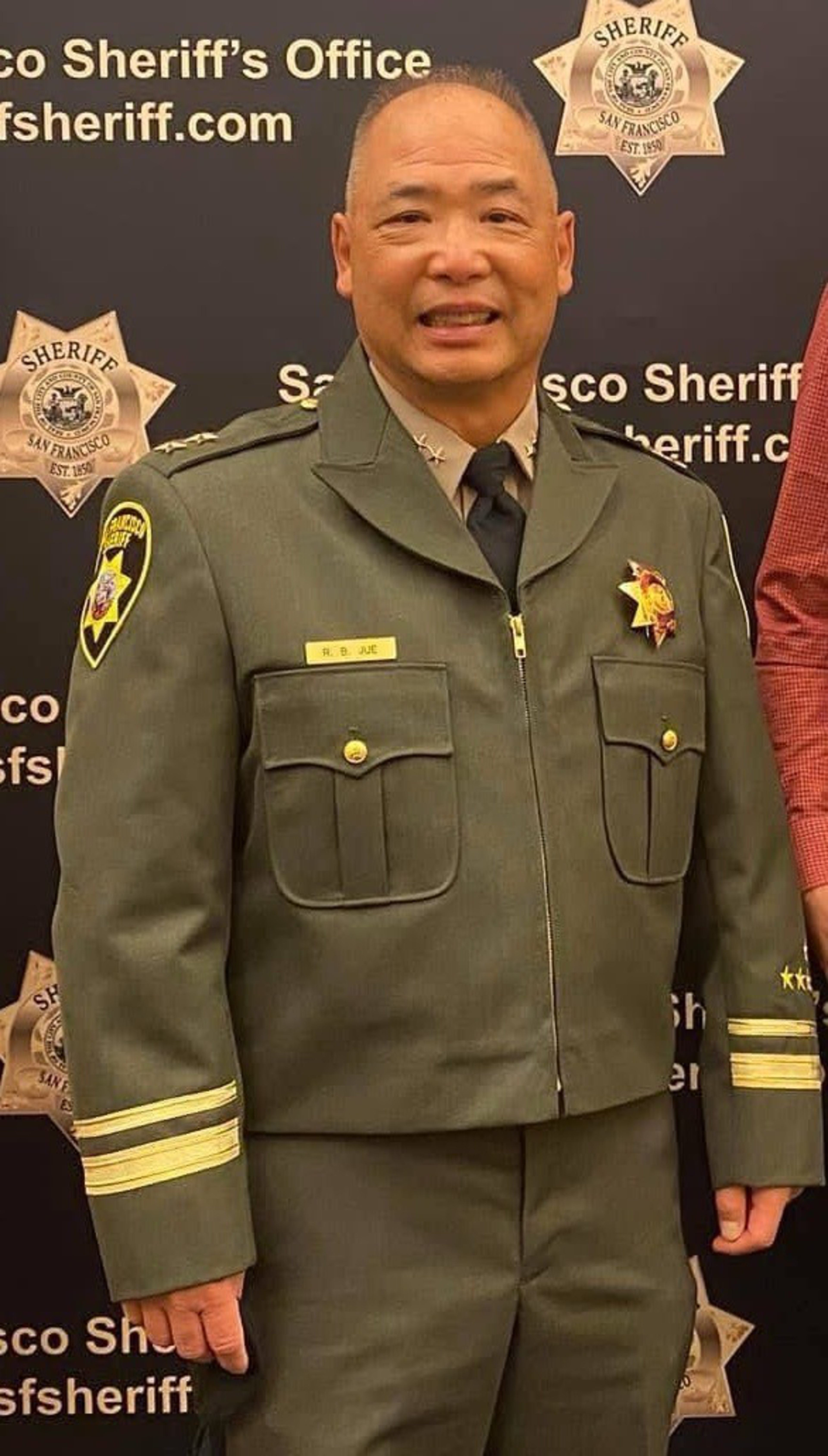A uniformed officer smiles, standing in front of a San Francisco Sheriff's Office backdrop. The uniform is dark with badges and a name tag.