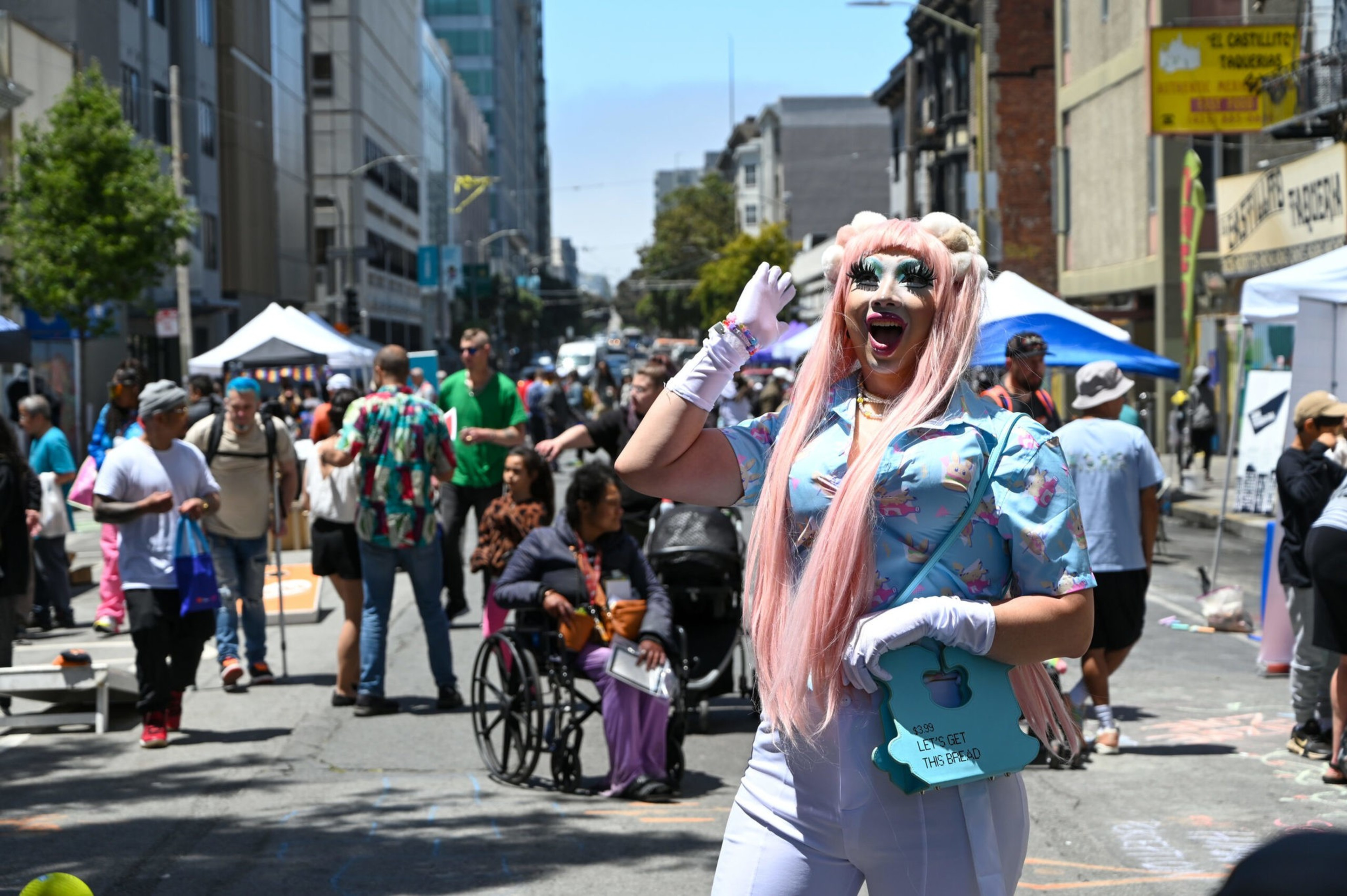 A person in vivid makeup and attire waves hello amidst a bustling street fair.