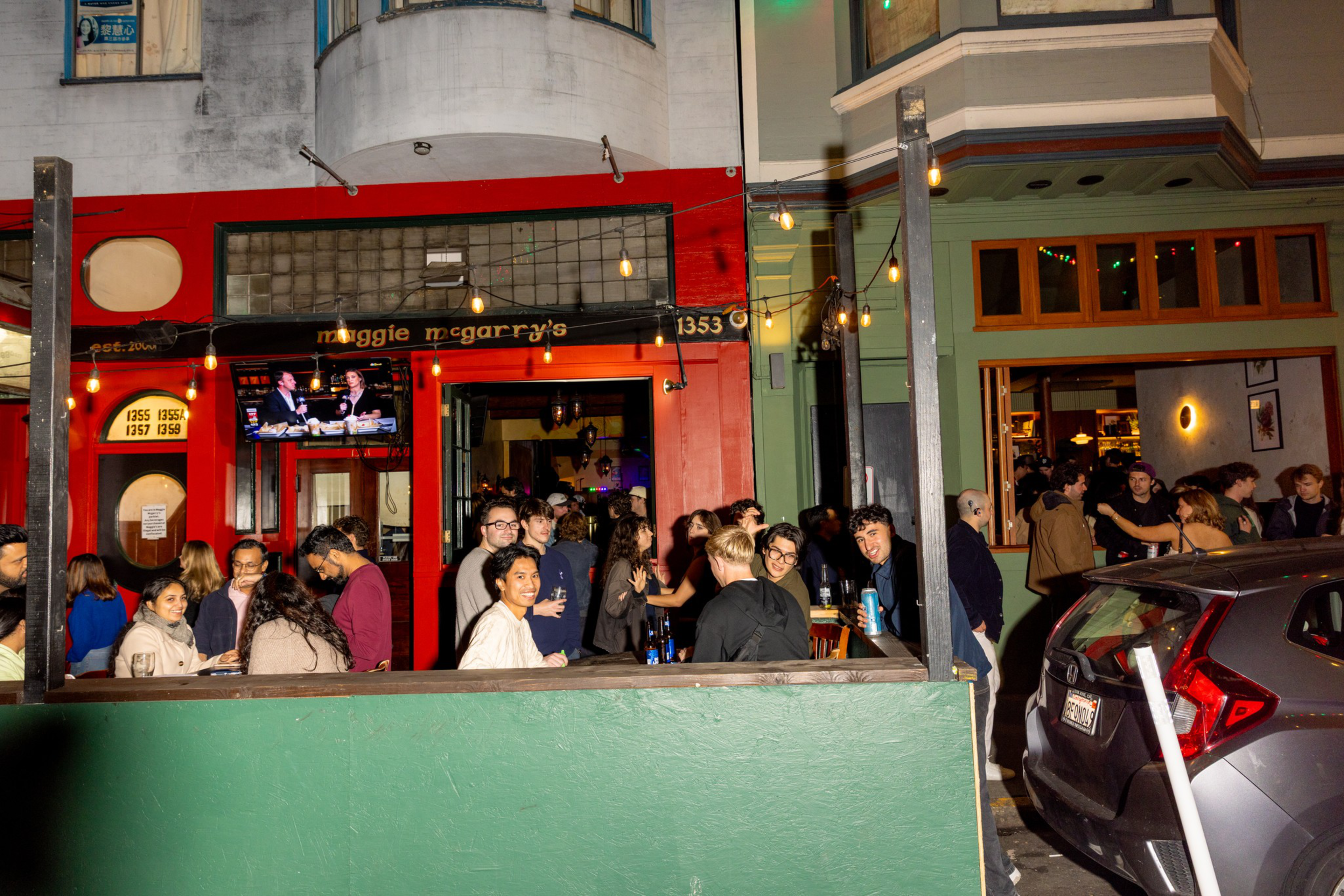 A lively crowd gathers outside and inside a brightly colored bar named &quot;Maggie McGarry&#x27;s&quot; with string lights and a TV showing a talk show.