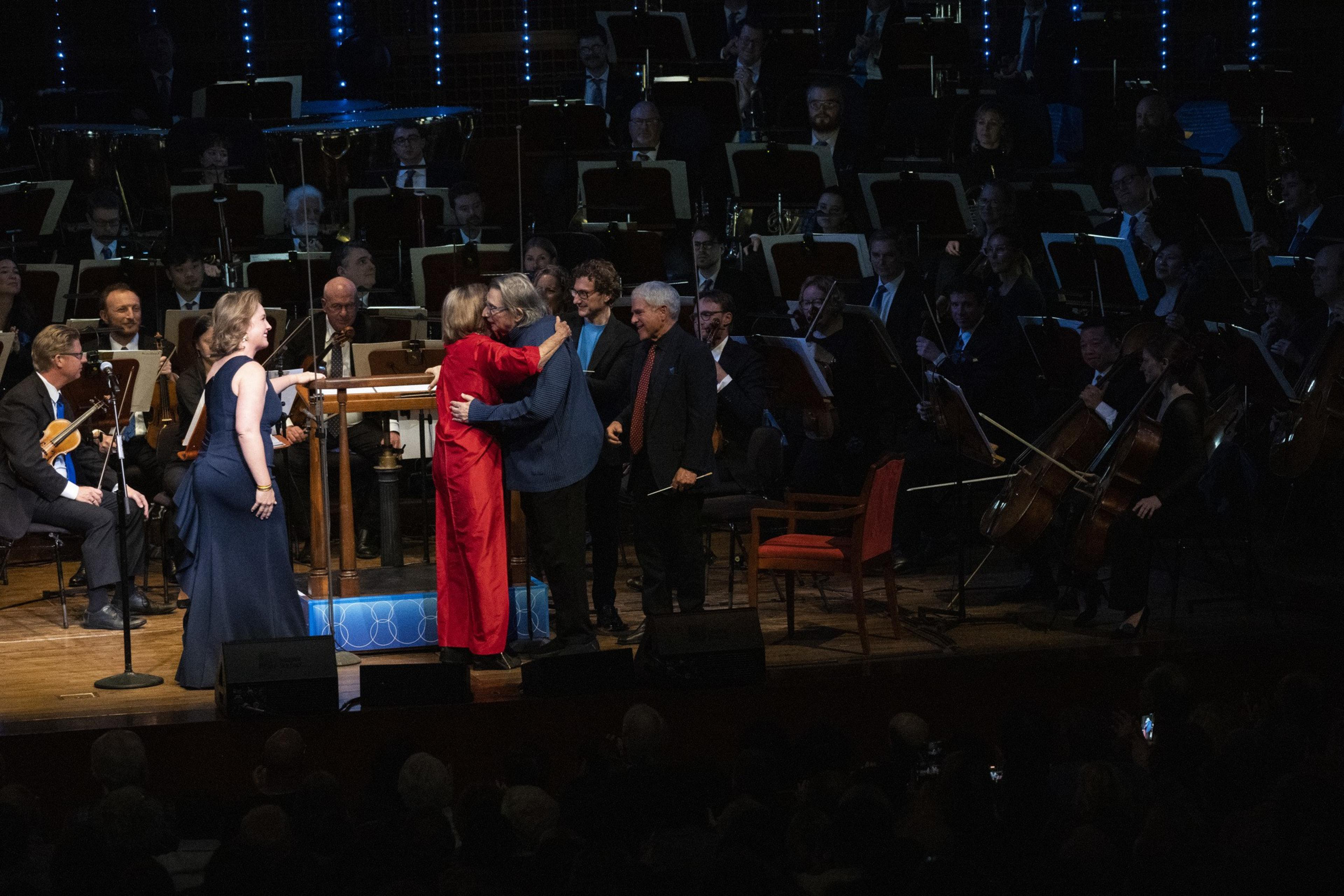 A group of people celebrates on stage in front of an orchestra. A woman in red embraces a man while others smile. Musicians with various instruments sit behind them.