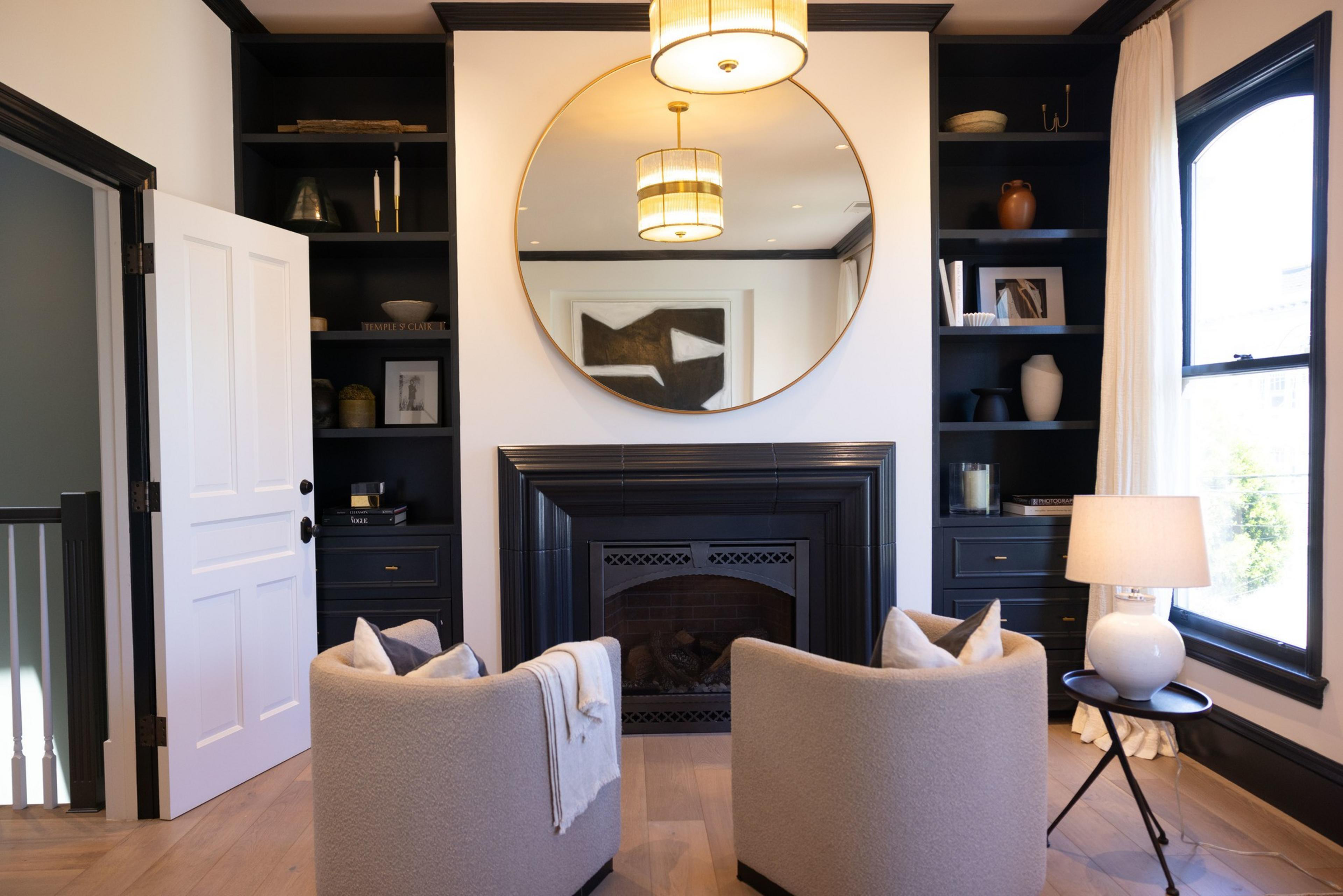 A cozy room with two beige chairs facing a black fireplace, above which hangs a large round mirror. Shelves with decor items, books, and lamps occupy both sides.