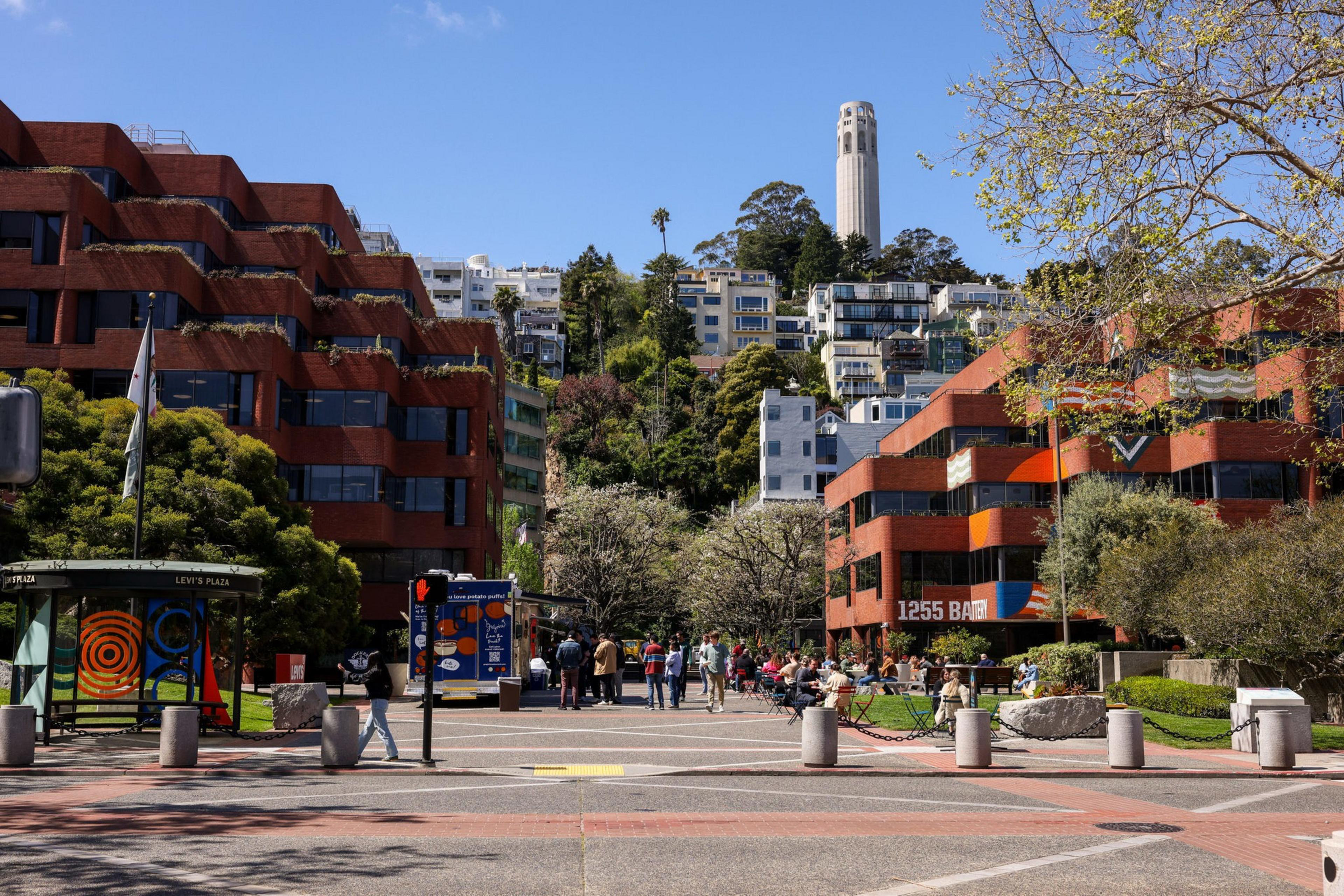 The image shows a bustling plaza with brick buildings and people relaxing. In the background, there's a hill with houses and a tall tower against a blue sky.