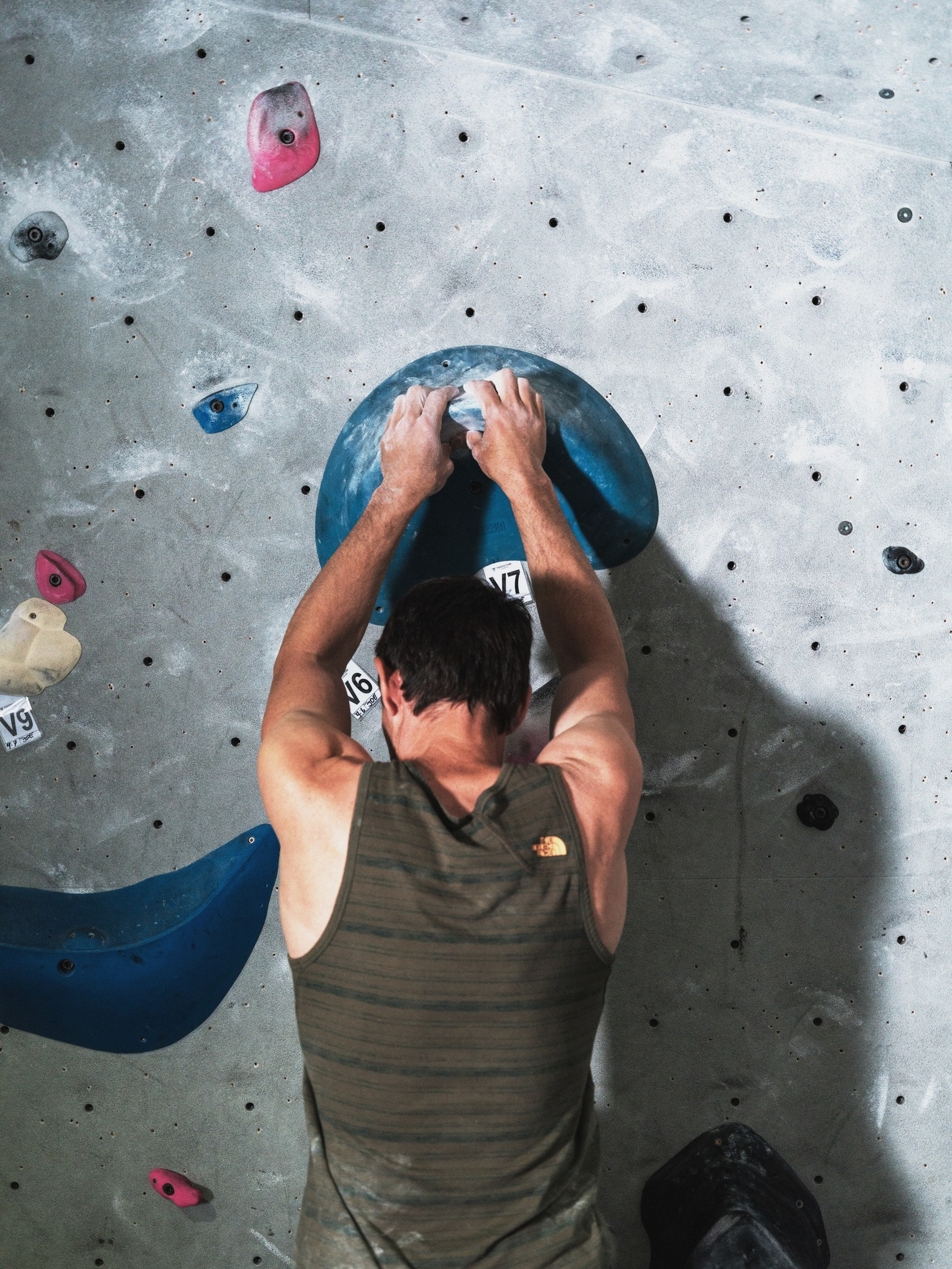 A person with short dark hair and a sleeveless shirt grips a large blue hold on an indoor climbing wall with various colored holds.