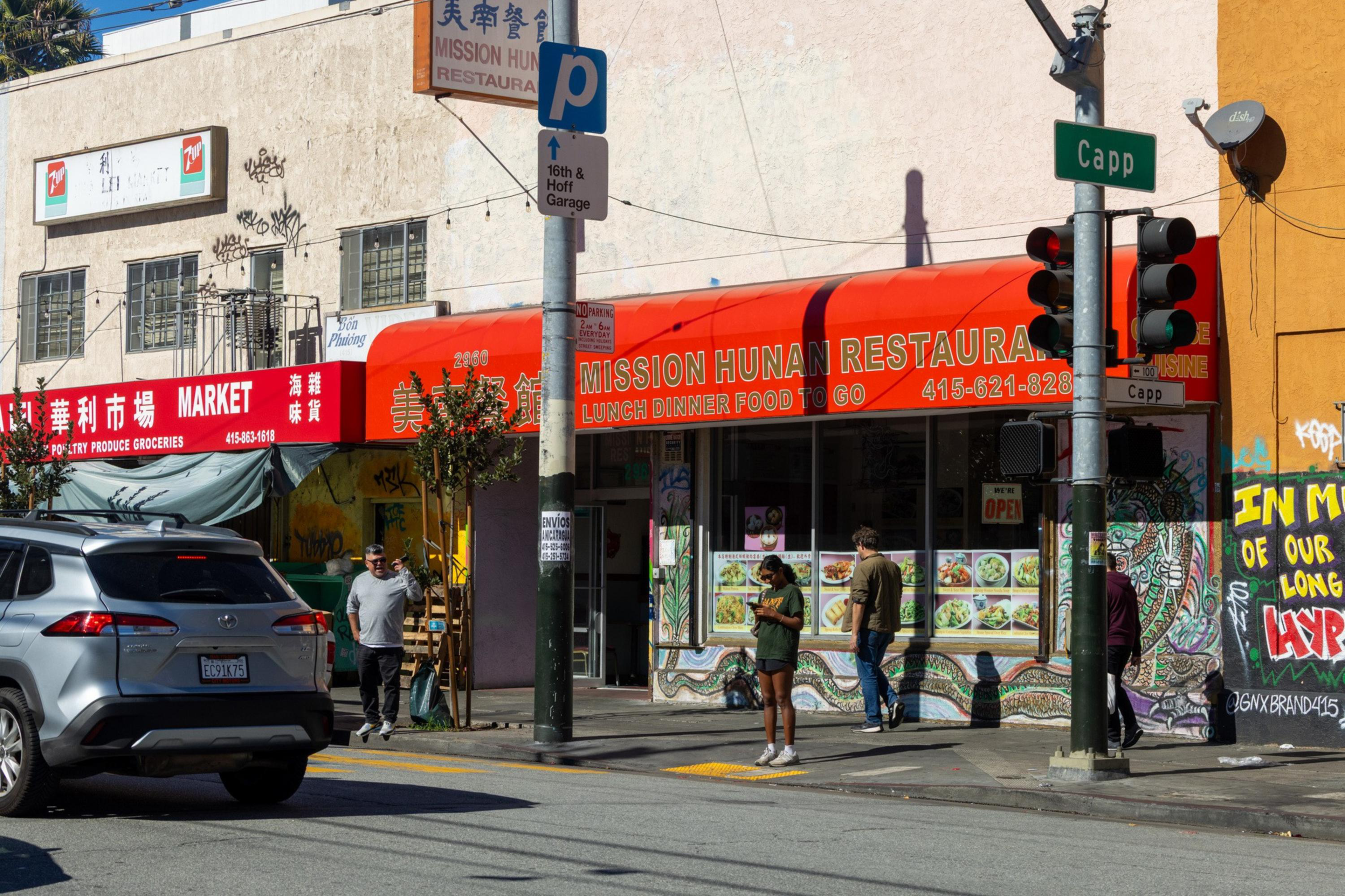 A red awning labeled “Mission Hunan Restaurant” covers a corner storefront with menu photos, a nearby market has red signage, and three people stand or walk by.