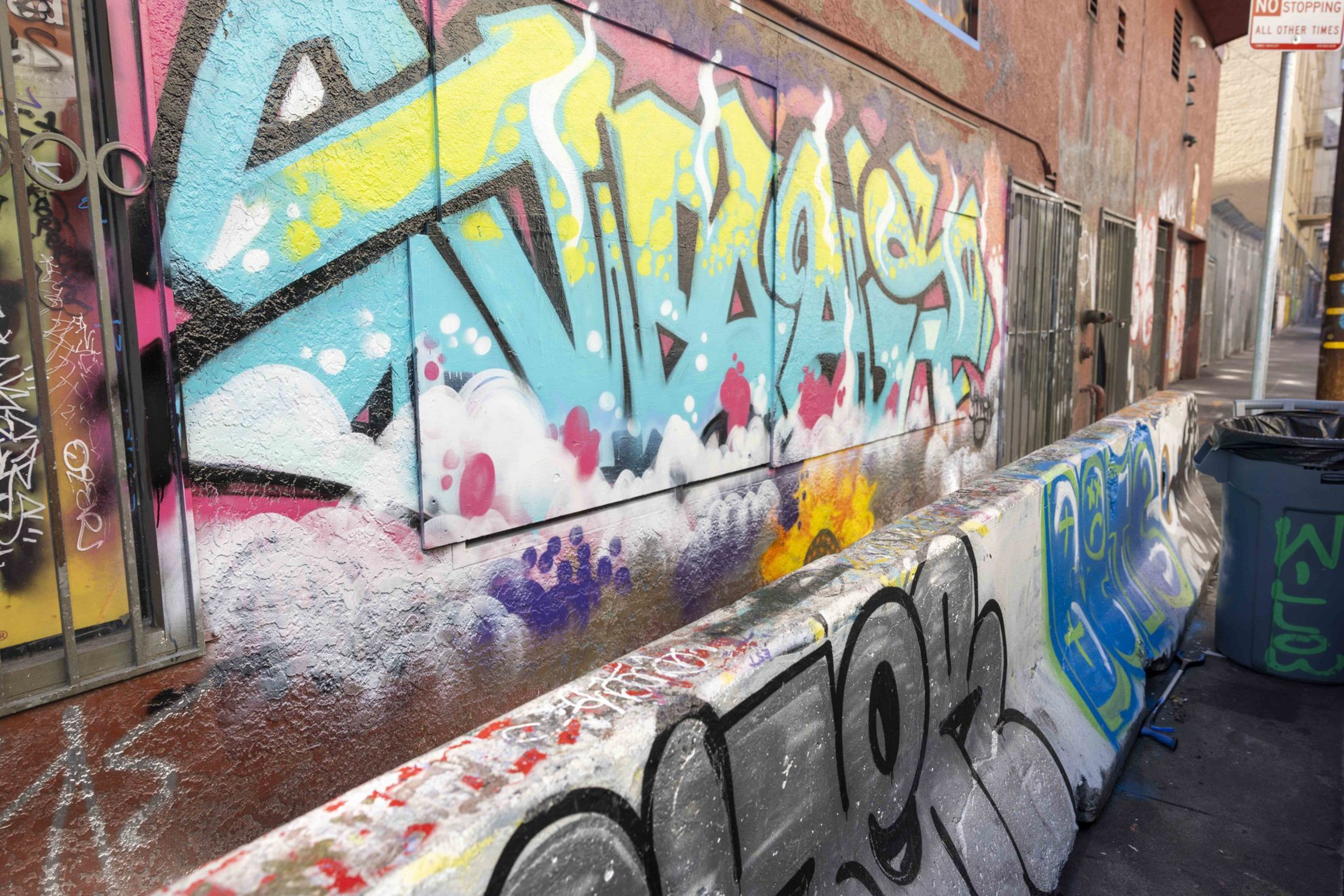 A colorful graffiti-covered alleyway with vibrant designs in blue, pink, and yellow. A trash bin and "No Stopping" sign are visible.