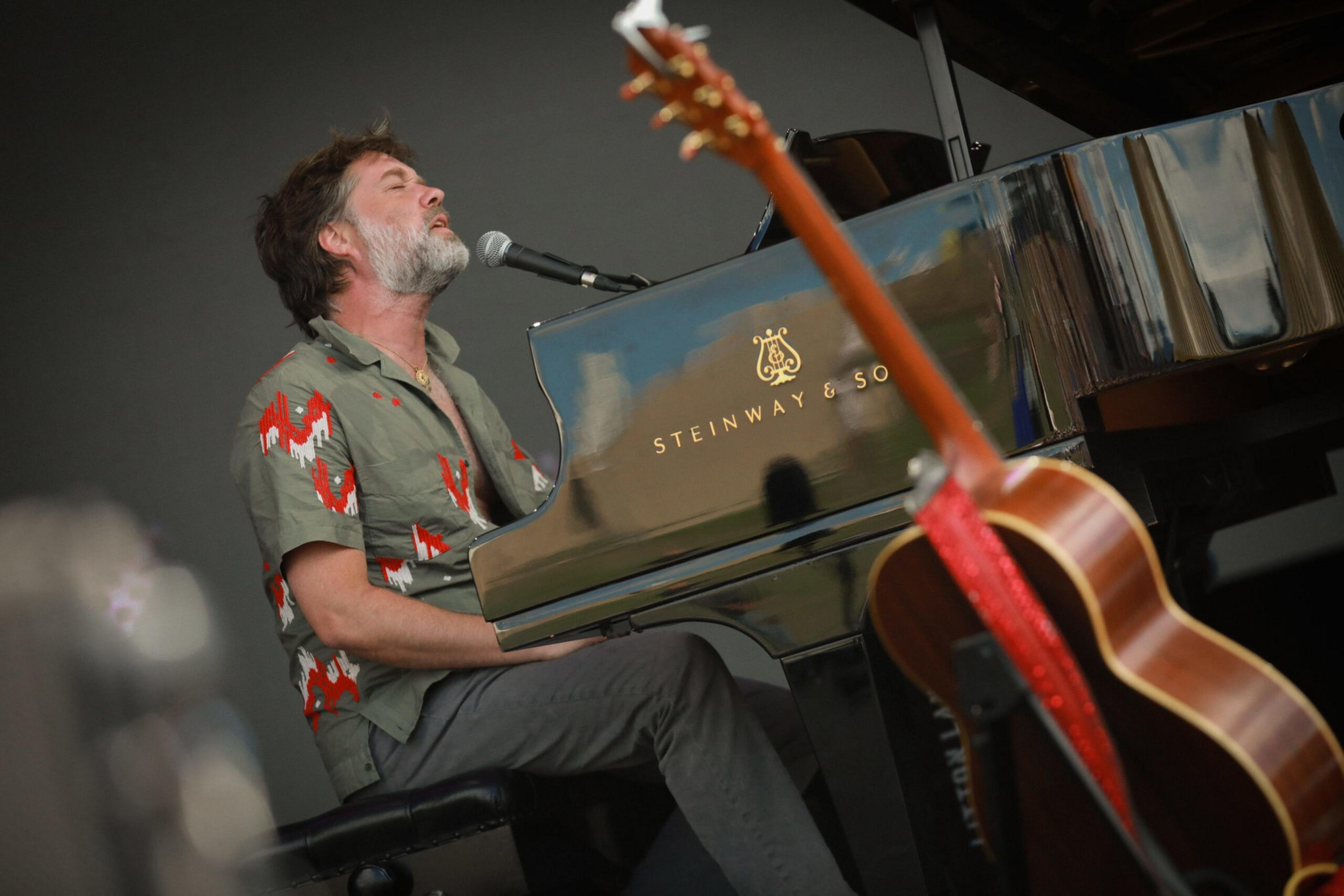 A man with a beard and patterned shirt sings into a microphone while playing a black Steinway & Sons piano, with a guitar in the foreground.