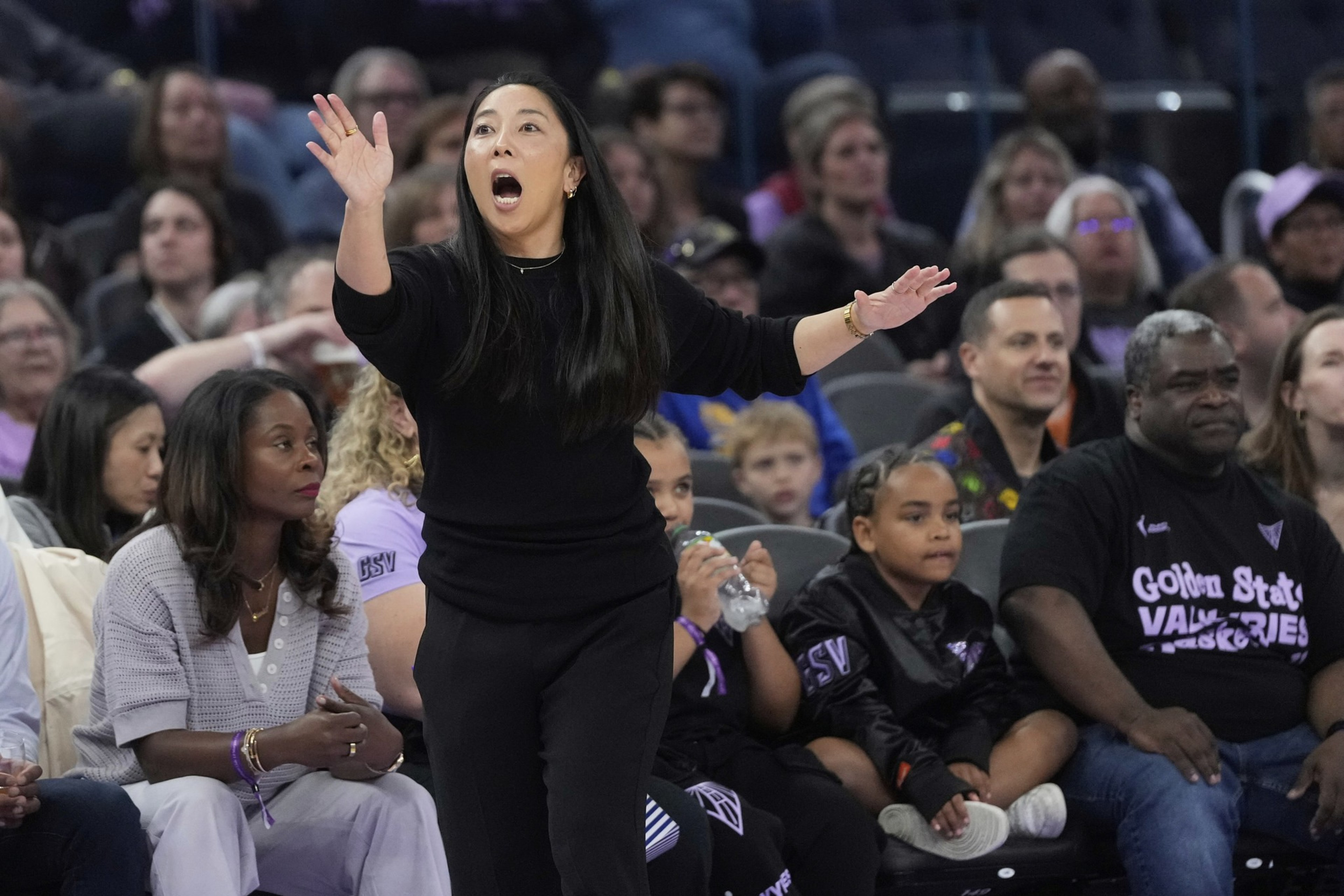 A woman in black passionately gestures from the sidelines at a sports event. Spectators fill the background, some focused on the action.