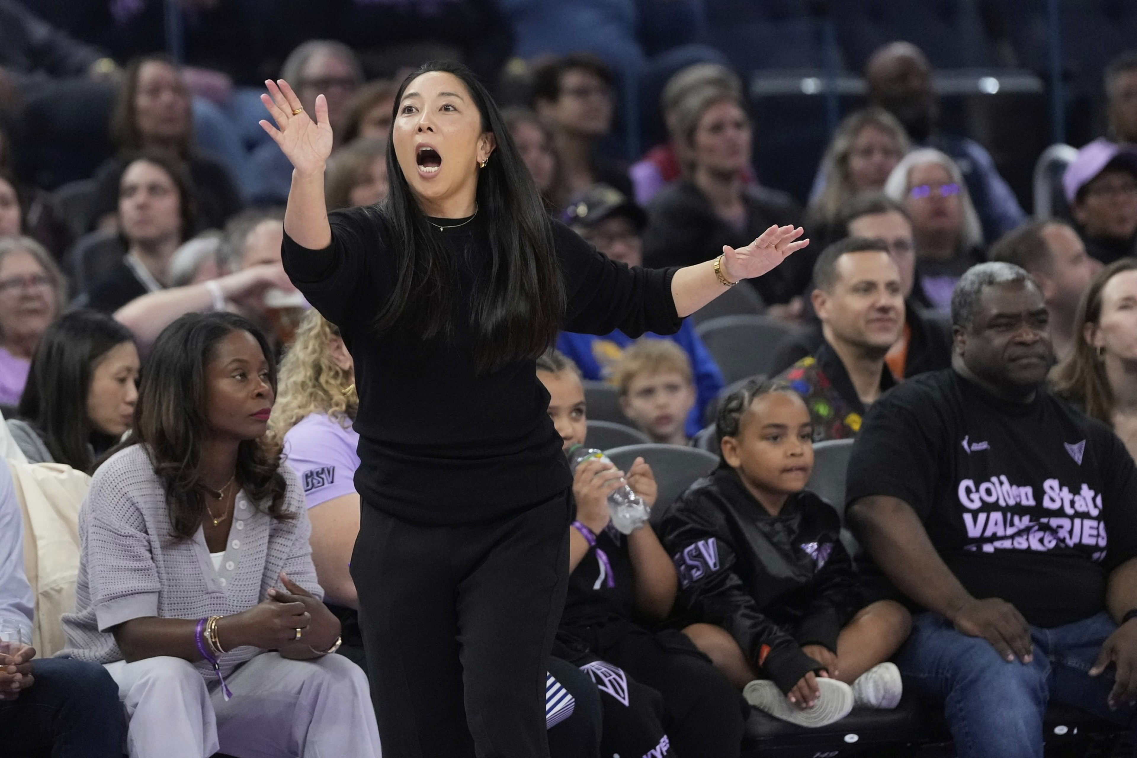A woman in black passionately gestures from the sidelines at a sports event. Spectators fill the background, some focused on the action.