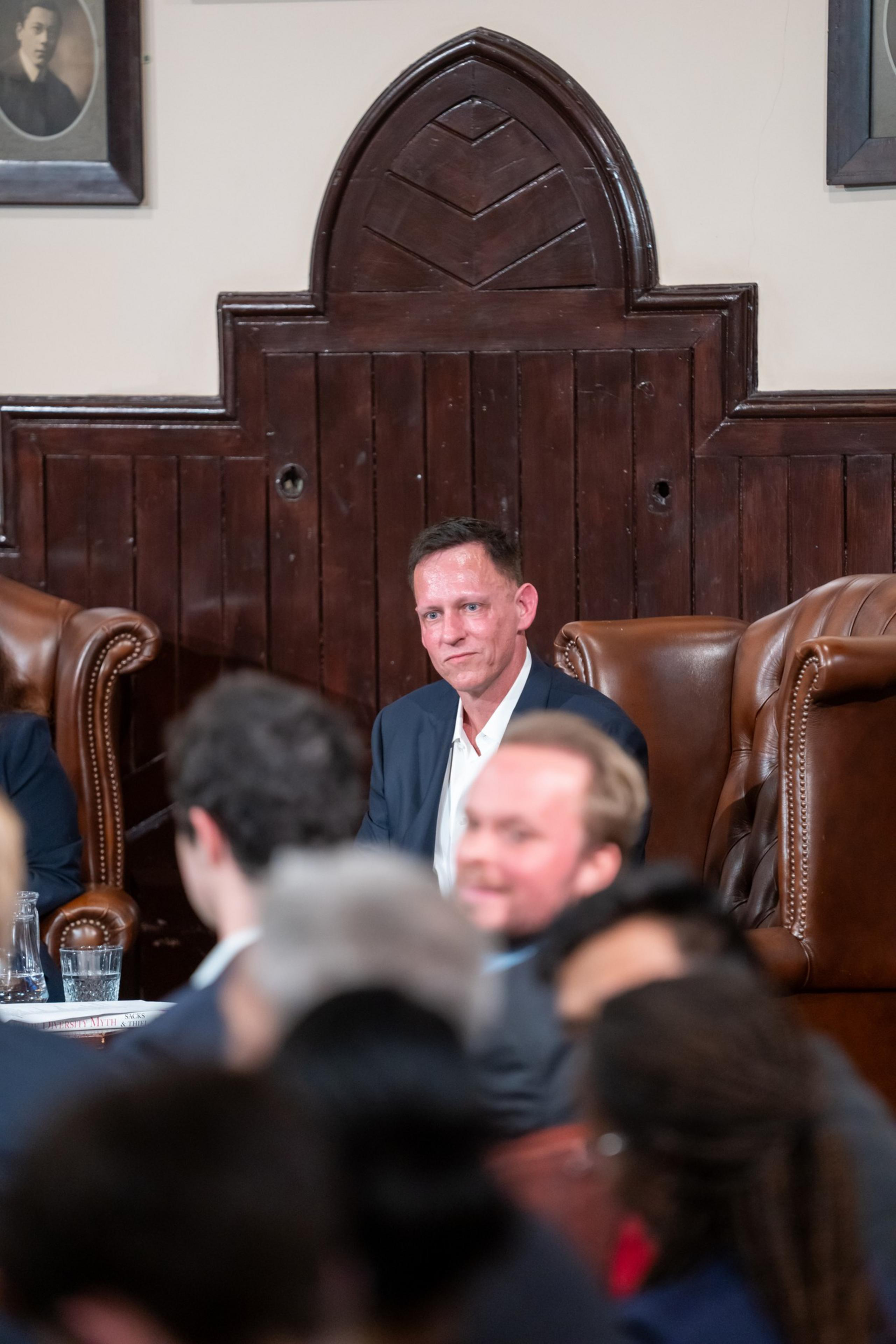 A man in a suit sits on a large brown leather chair against a dark wooden paneled wall, surrounded by blurred people in the foreground.