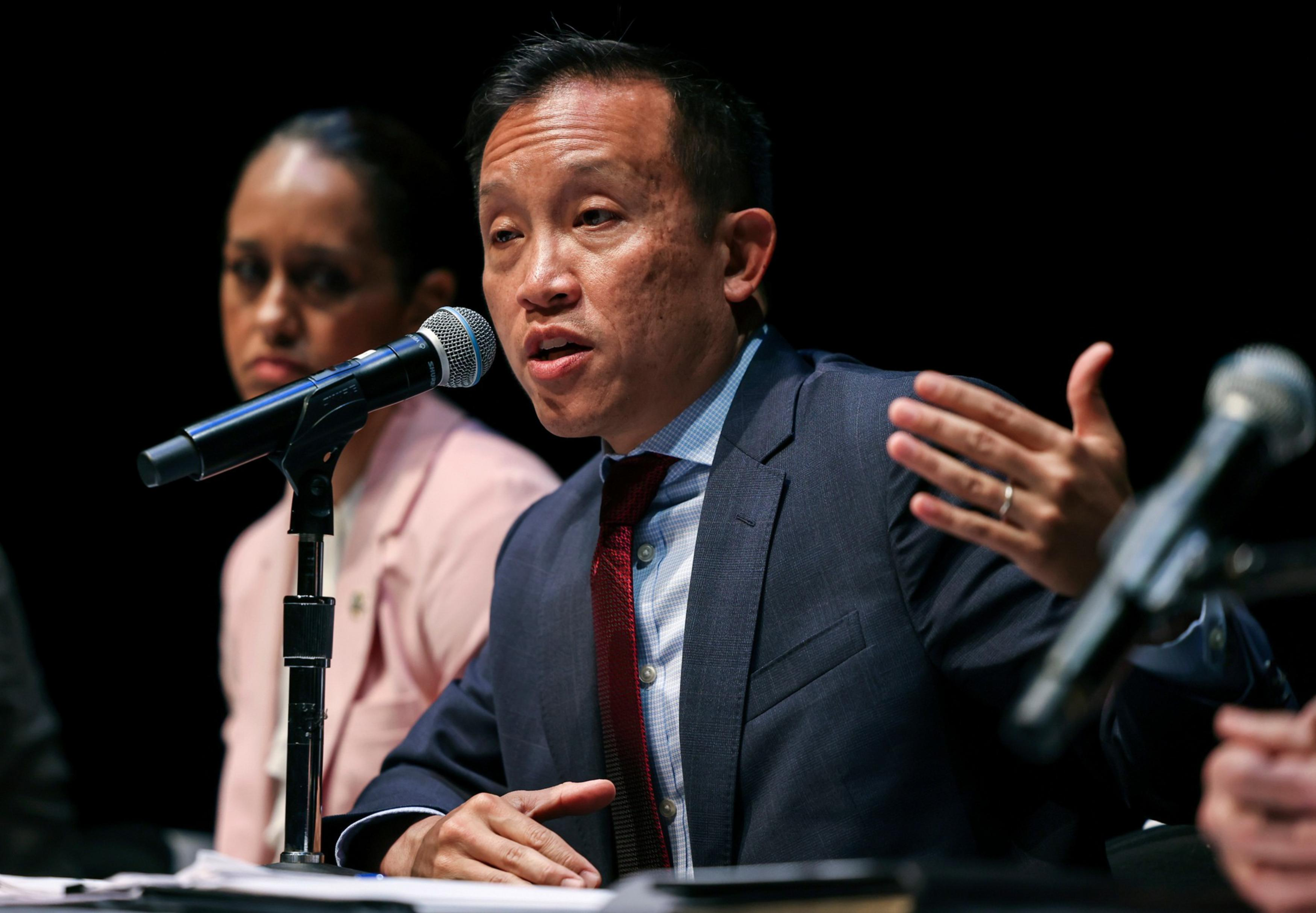 A man in a suit speaks into a microphone with one hand raised, while a woman in a pink jacket listens attentively behind him.