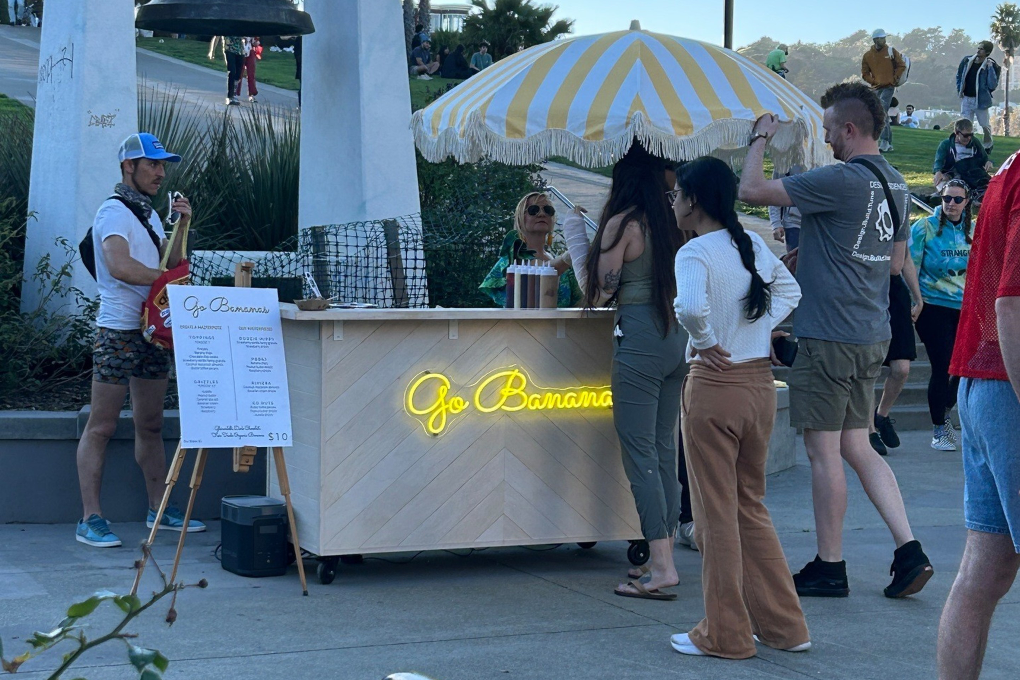 A small food stand named "Go Banana" with a yellow-striped umbrella serves customers near a large outdoor bell and palm trees on a sunny day.