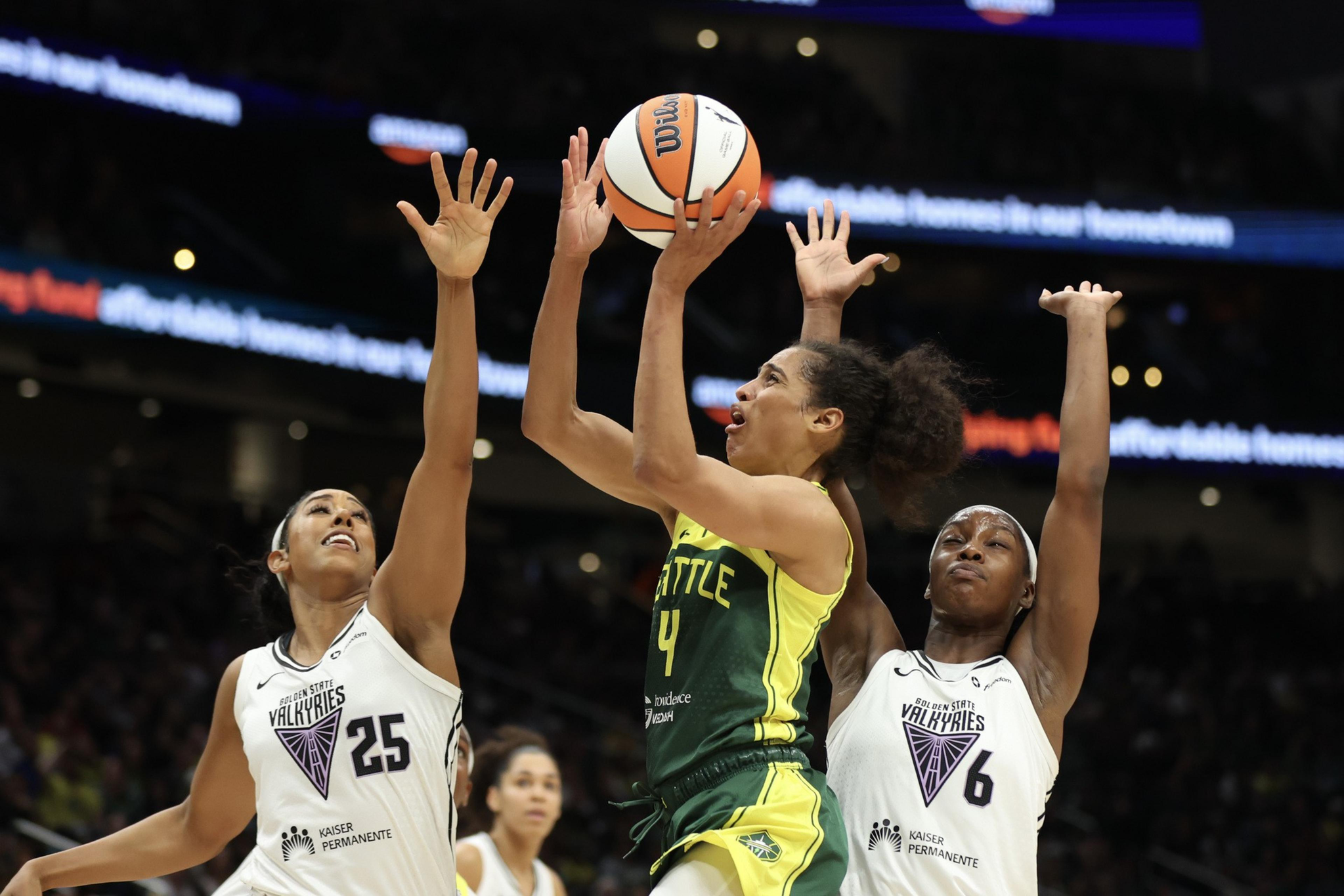 A Seattle player wearing green and yellow jumps to shoot a basketball while two opposing players in white attempt to block the shot.