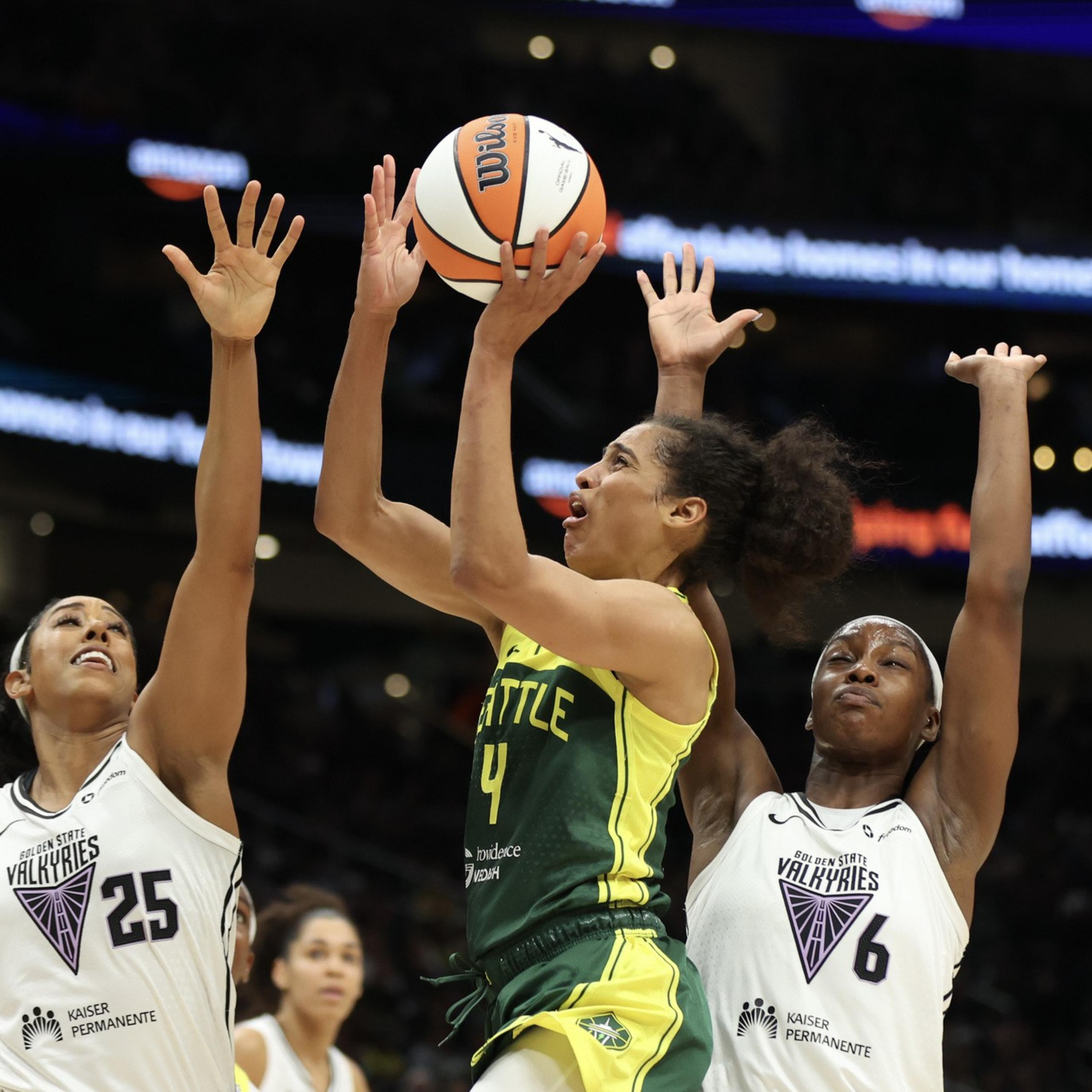A Seattle player wearing green and yellow jumps to shoot a basketball while two opposing players in white attempt to block the shot.