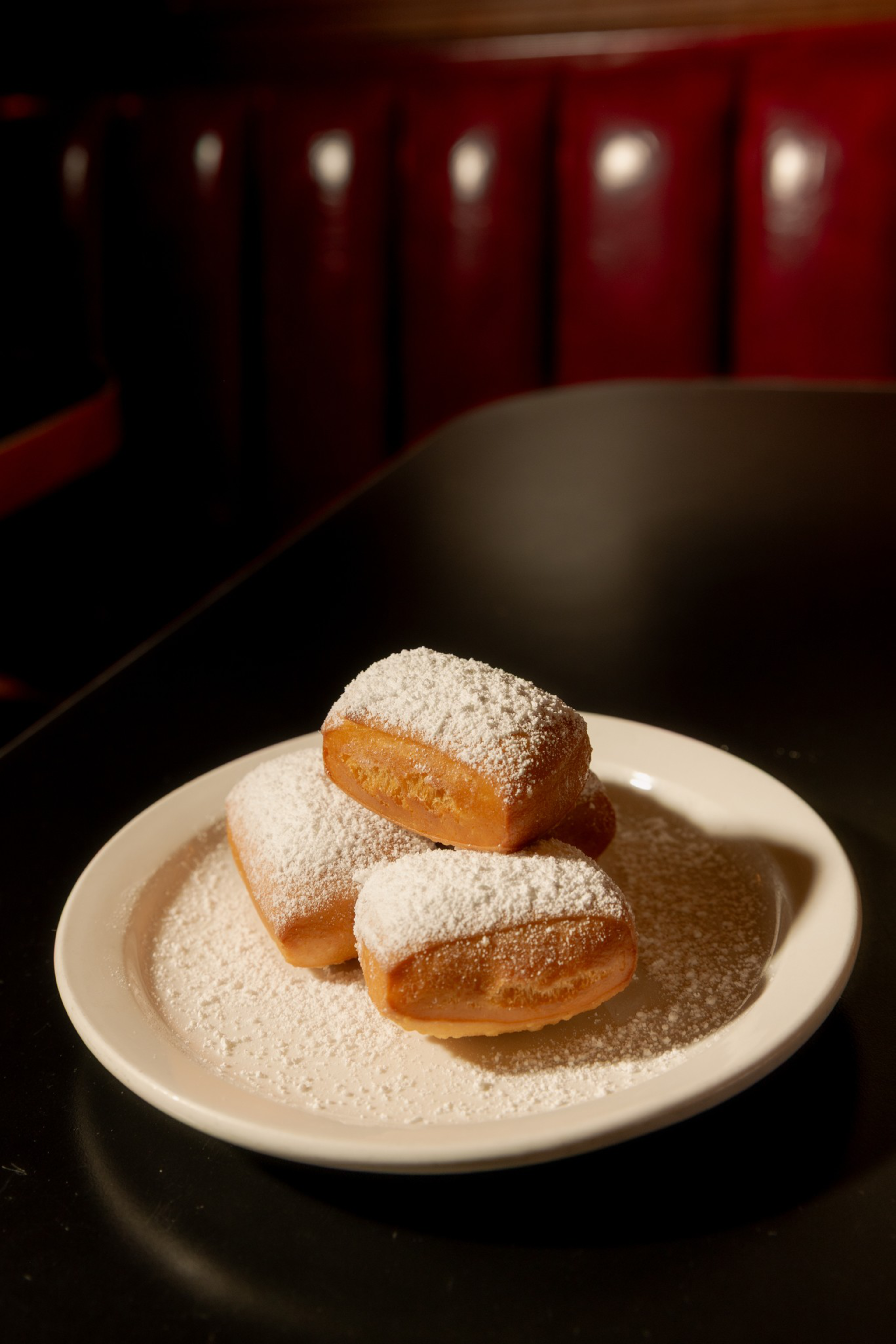Three rectangular beignets covered in powdered sugar sit stacked on a white plate on a dark table with a red cushioned booth in the background.