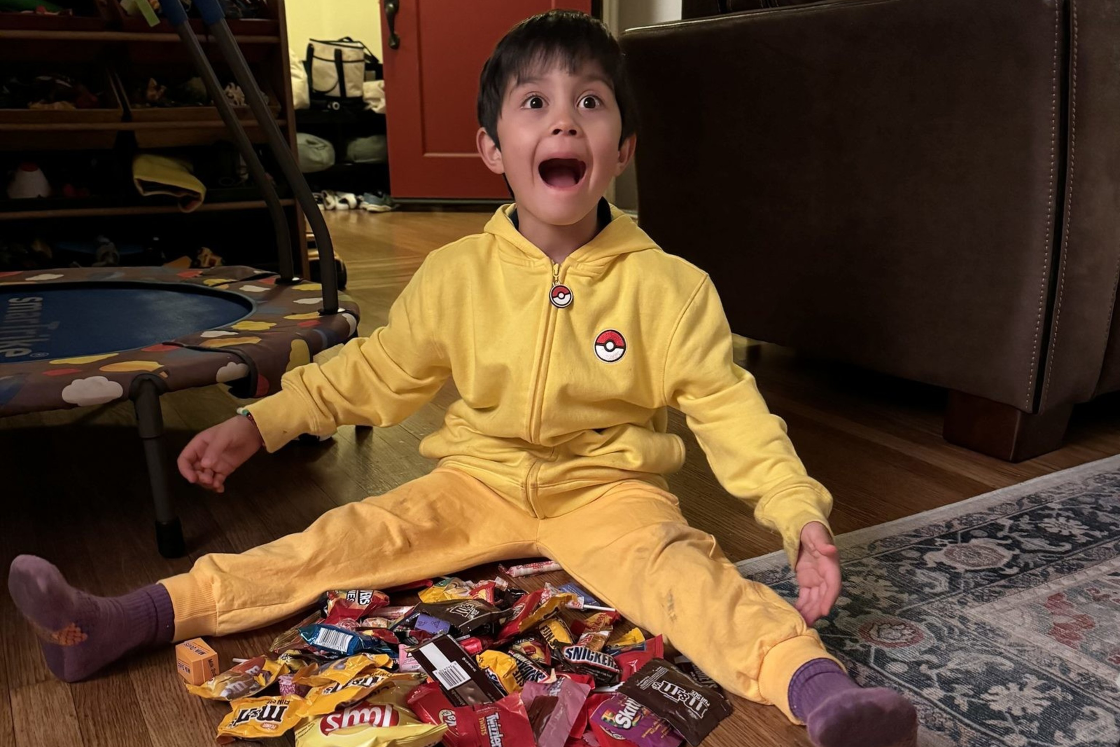 A kid with brown hair wearing a yellow zip-up sweater and yellow pants sits on the ground with candy out in front of him.