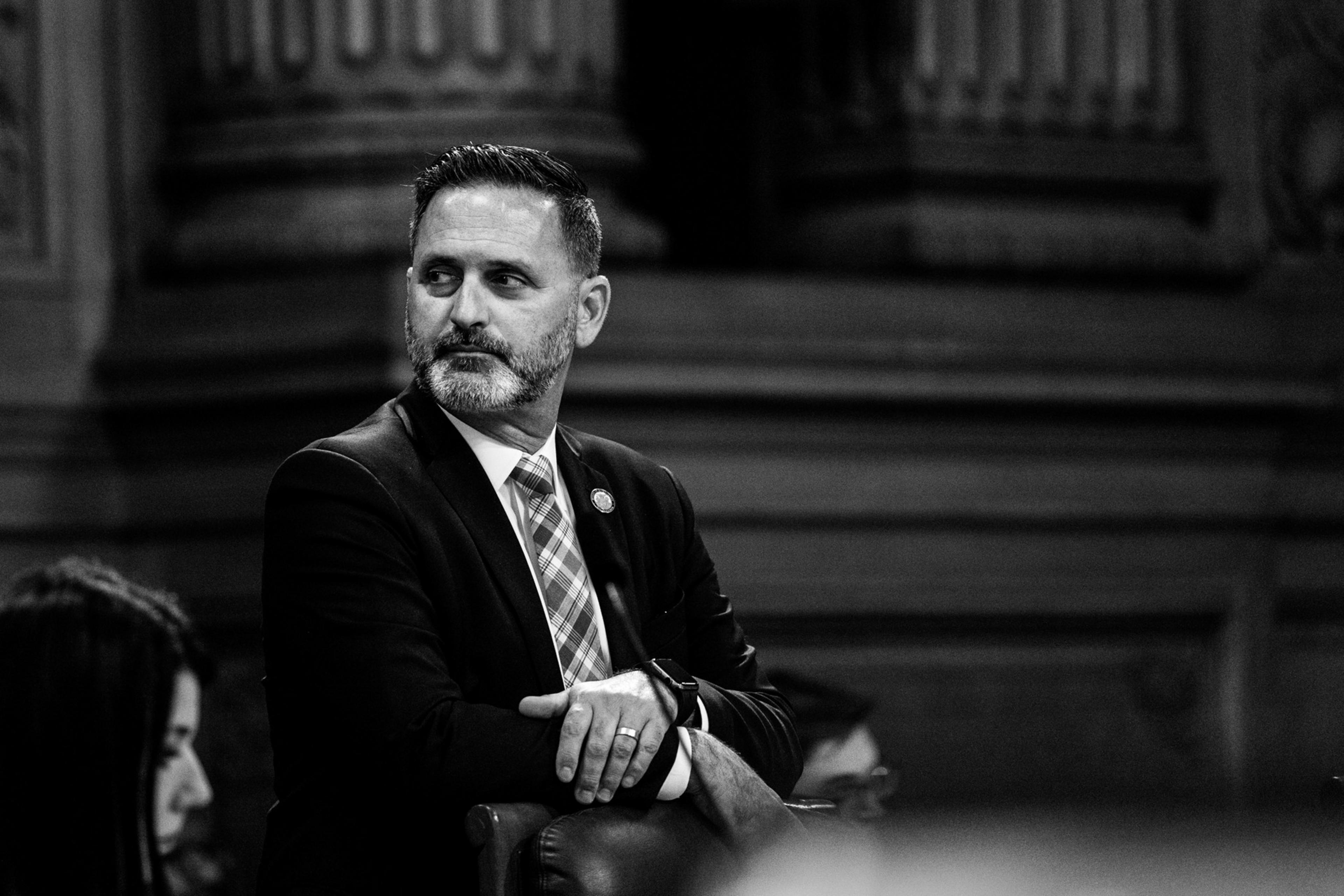 A man in a suit looks to the side, seated with interlocked hands in a formal setting. Black and white tones enhance the solemn mood.
