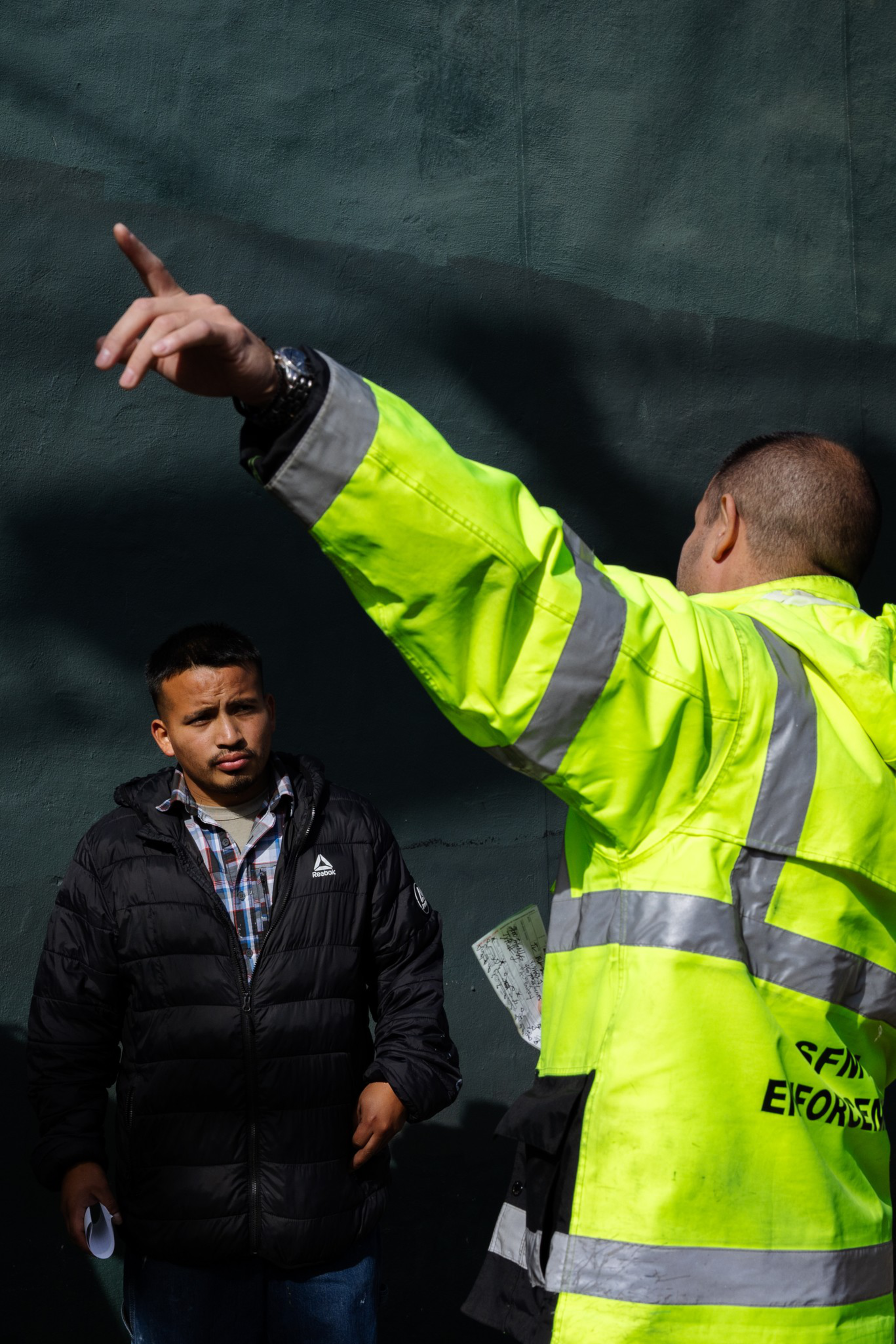 A man in a bright yellow reflective jacket points while another man in a black jacket with a plaid shirt underneath watches attentively.