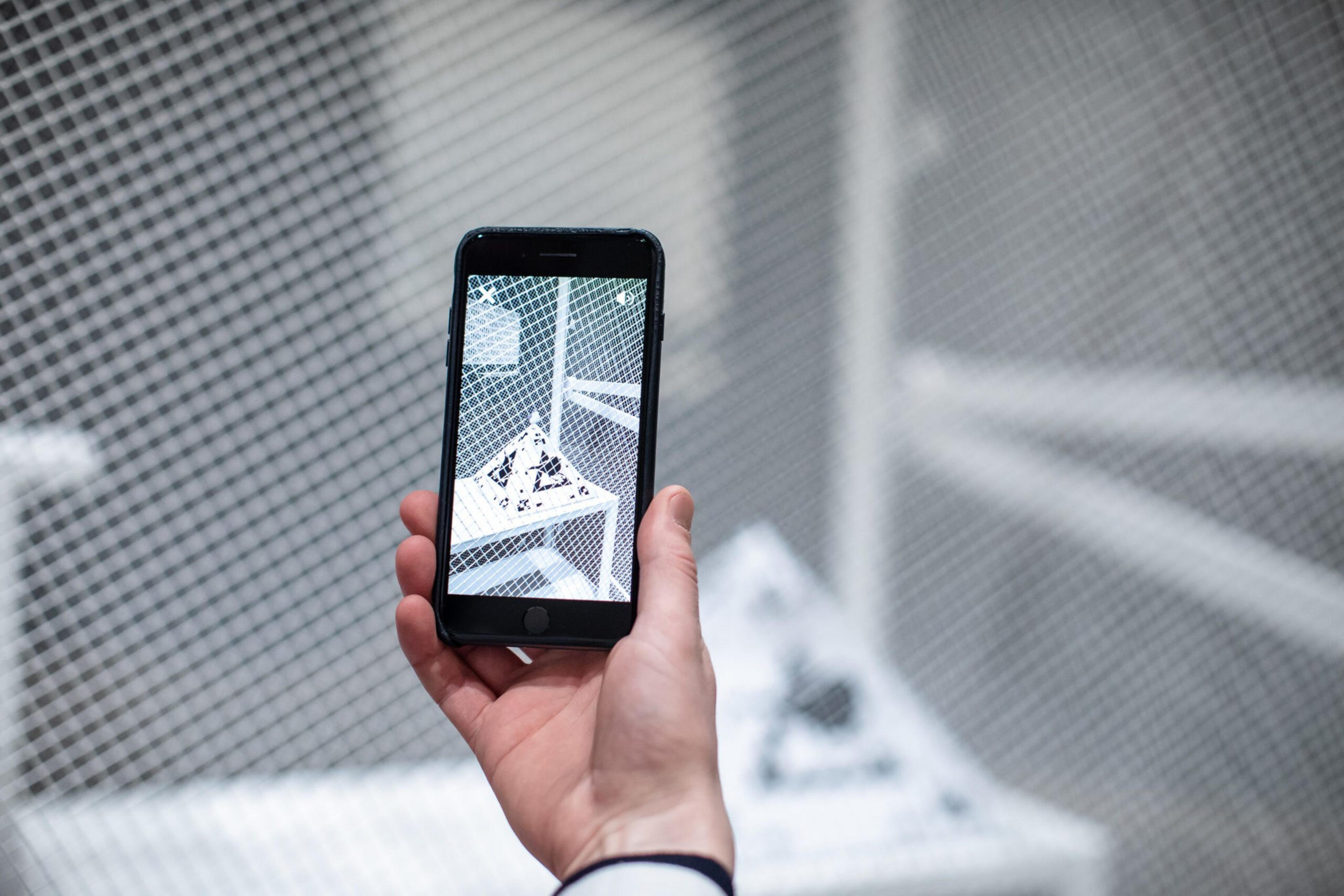 A hand holds a smartphone capturing a close-up of a white mesh structure with geometric shapes inside, framed by the same mesh in the background.