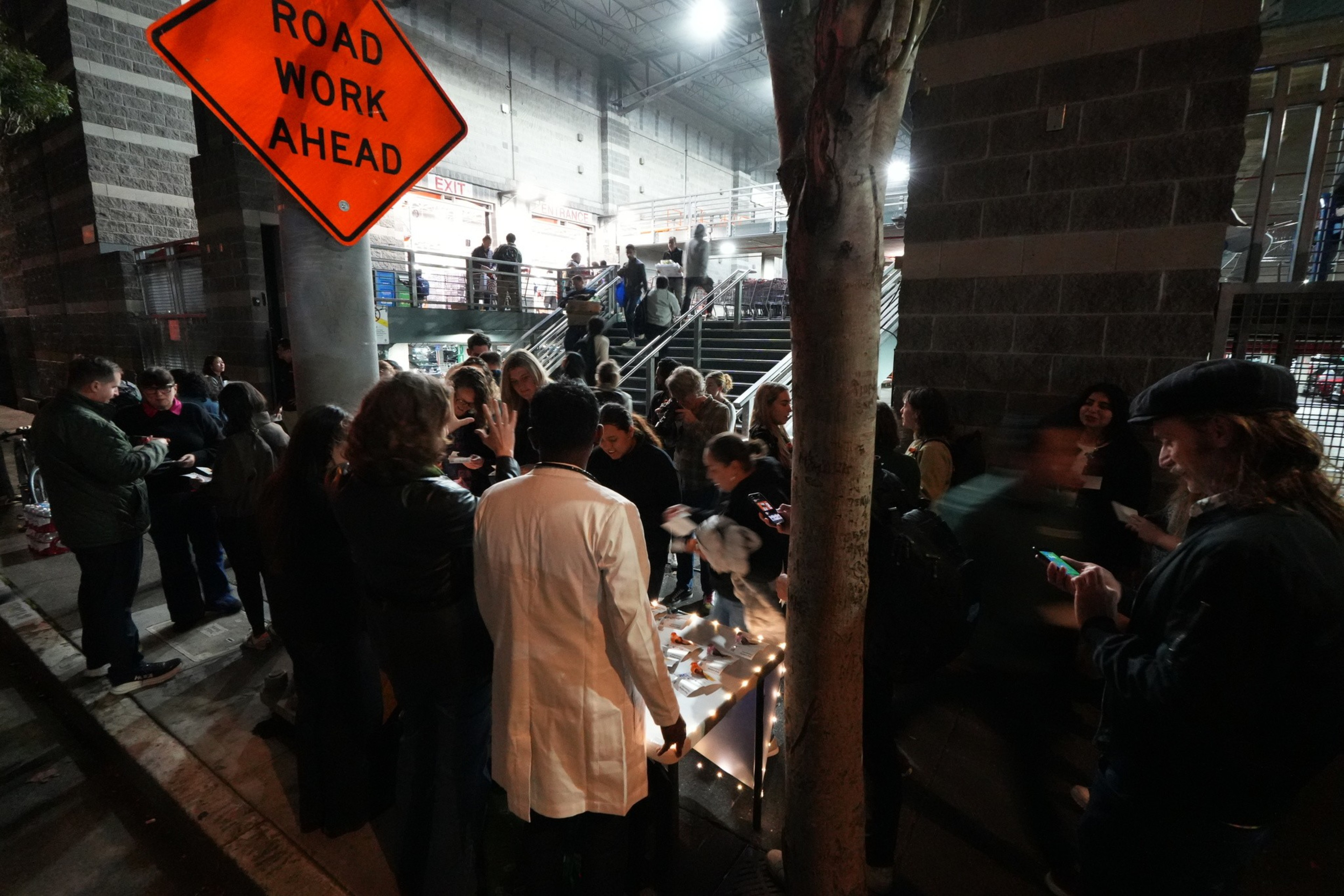 A group of people gathers around a small illuminated memorial or display on a sidewalk near stairs, under a “Road Work Ahead” sign at night.