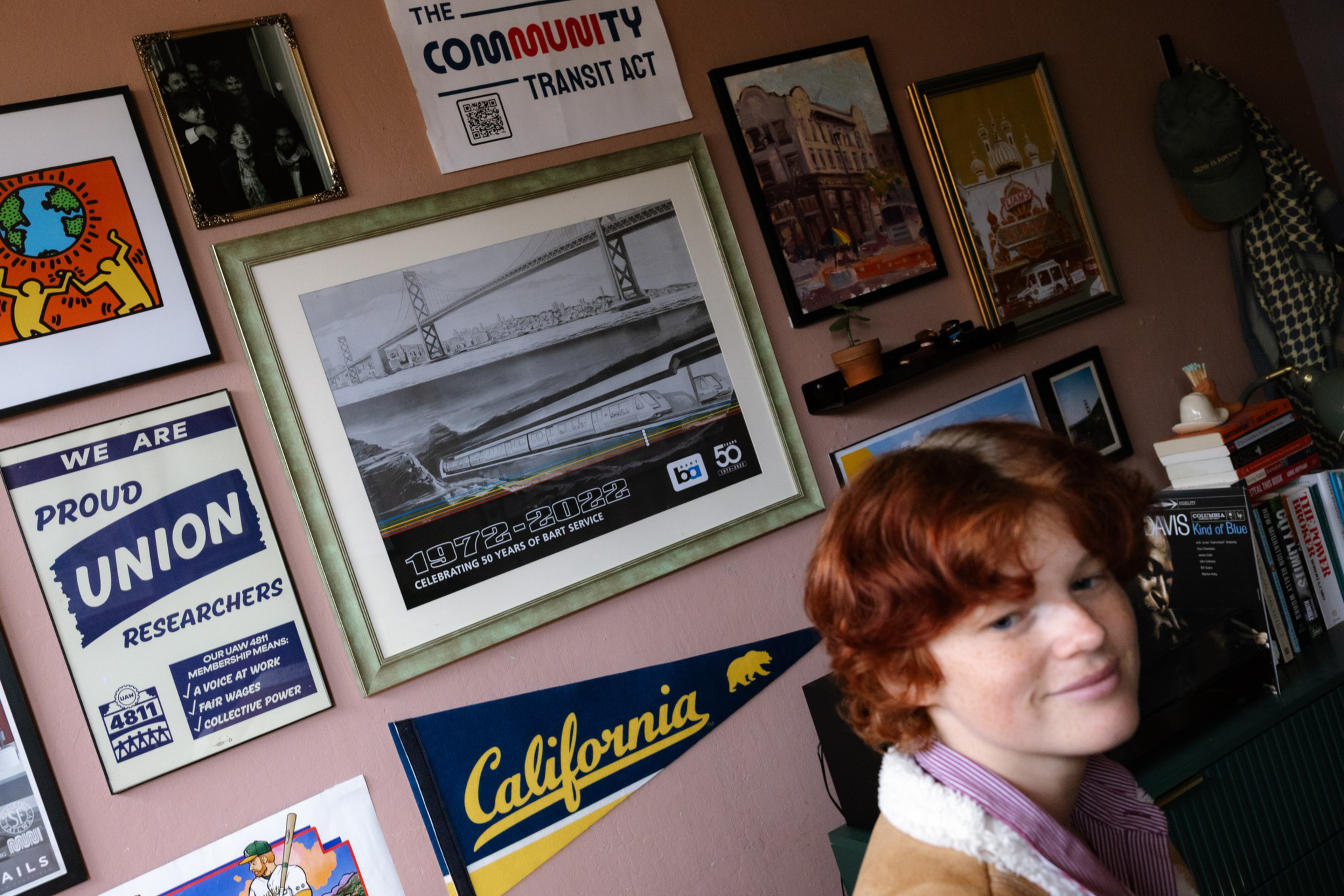 A young person with curly red hair sits in a room filled with framed posters, union signs, a California pennant, and books on shelves.