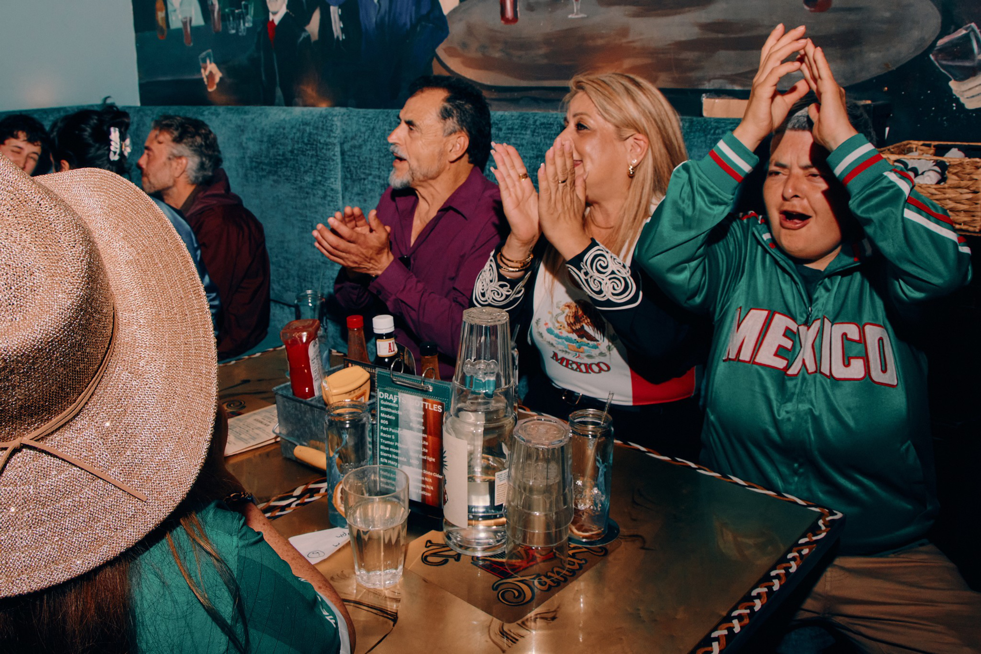 People sit at a table applauding and cheering; one person wears a green “MEXICO” jacket, and another has a large straw hat. Various drinks and condiments are on the table.