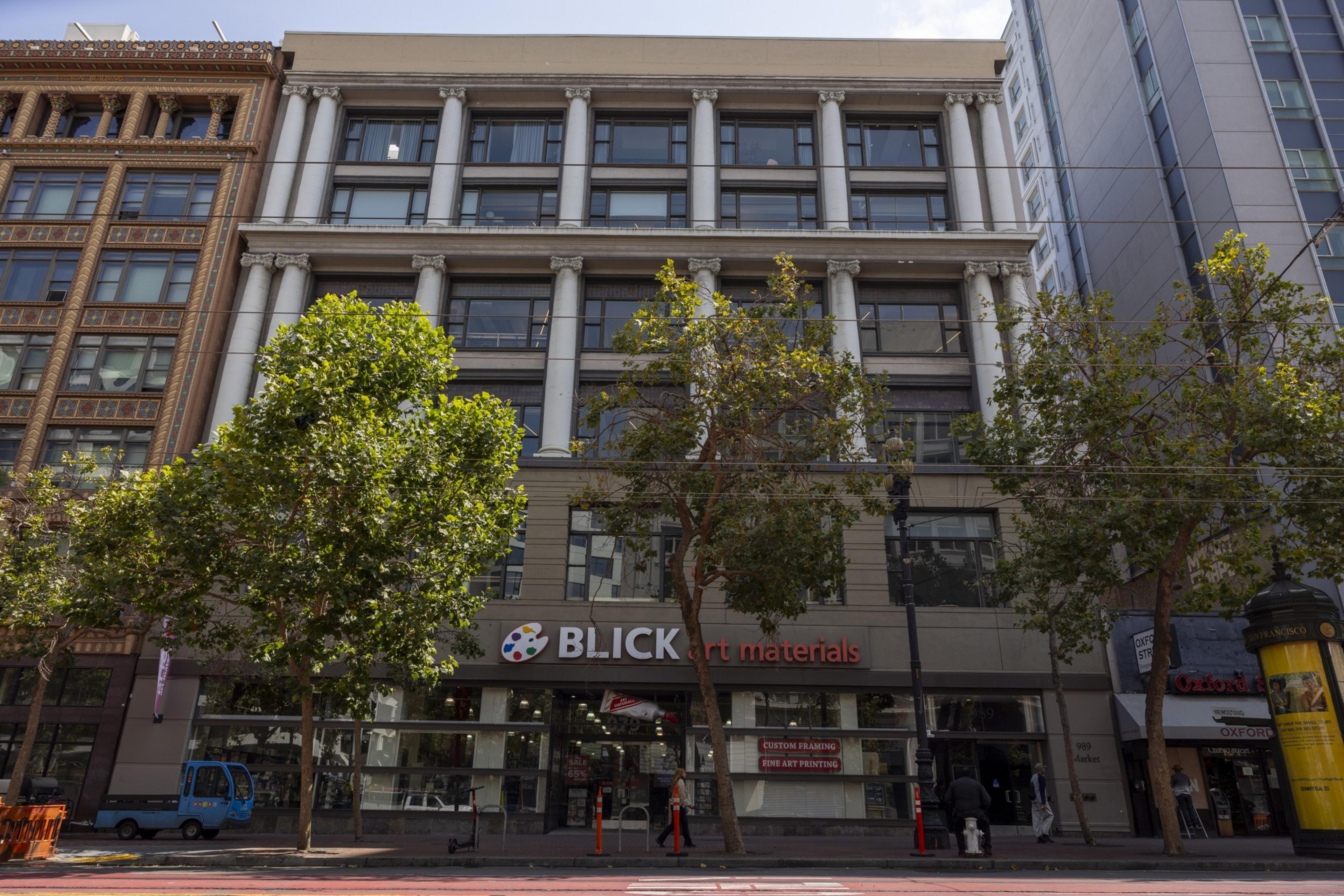The image shows a multi-story building with a store named &quot;Blick Art Materials&quot; on the ground floor, surrounded by trees and other adjacent buildings on a busy street.