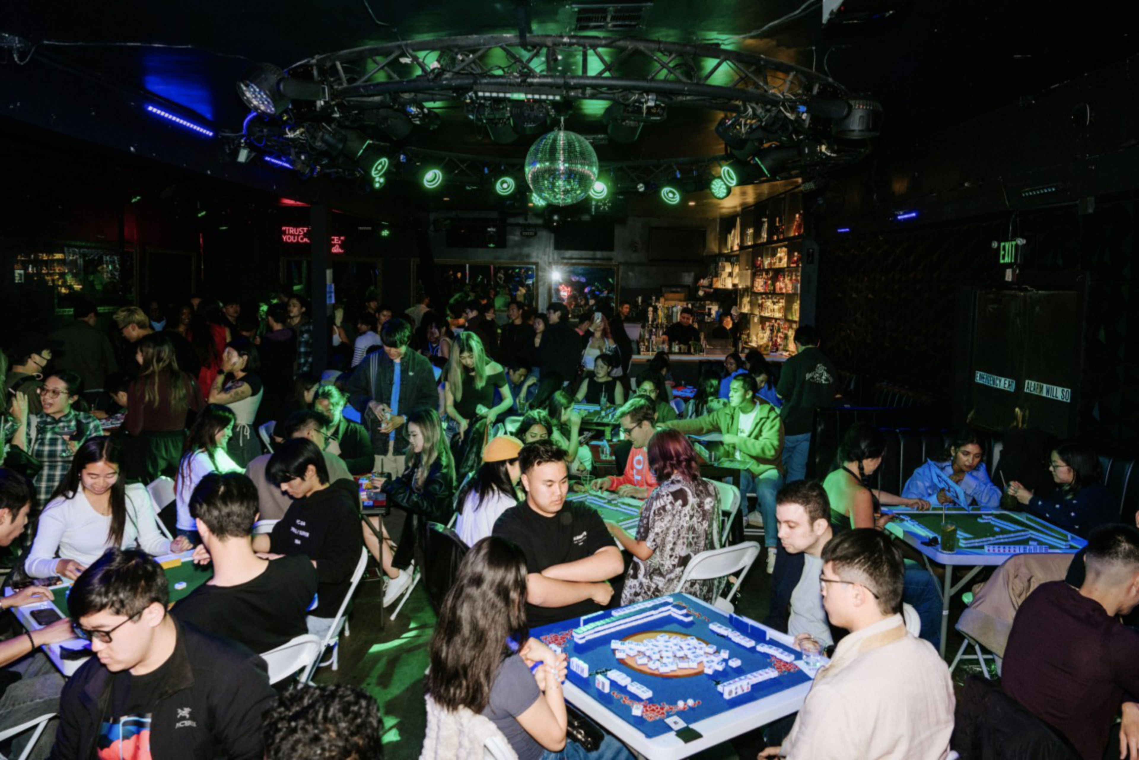 A crowded, dimly lit room with green lighting where people sit at tables playing mahjong under a disco ball near a stocked bar.