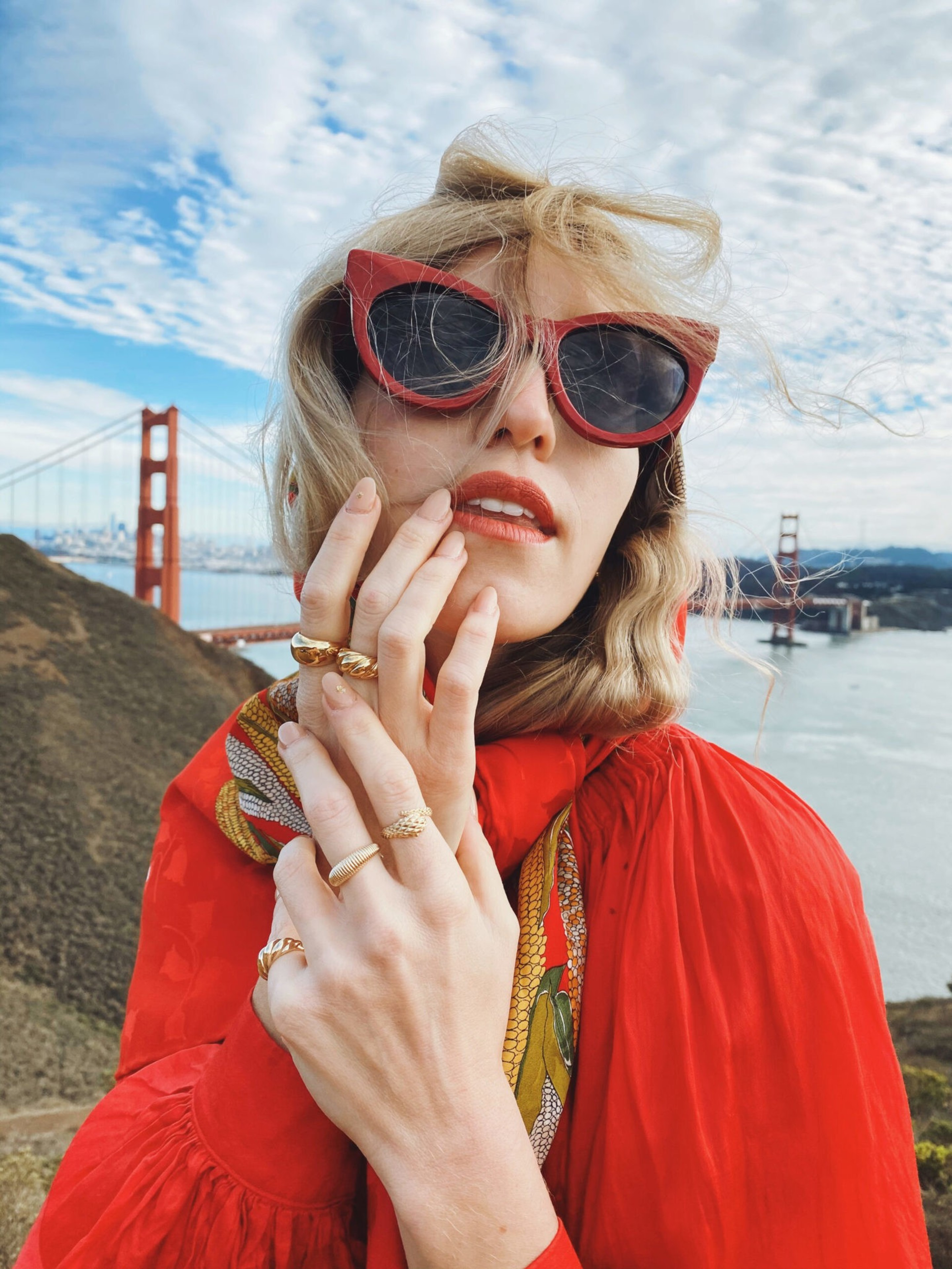 A woman wearing bold red sunglasses, lipstick, a red outfit, and gold rings poses near the Golden Gate Bridge with wind-tousled hair and a colorful scarf.