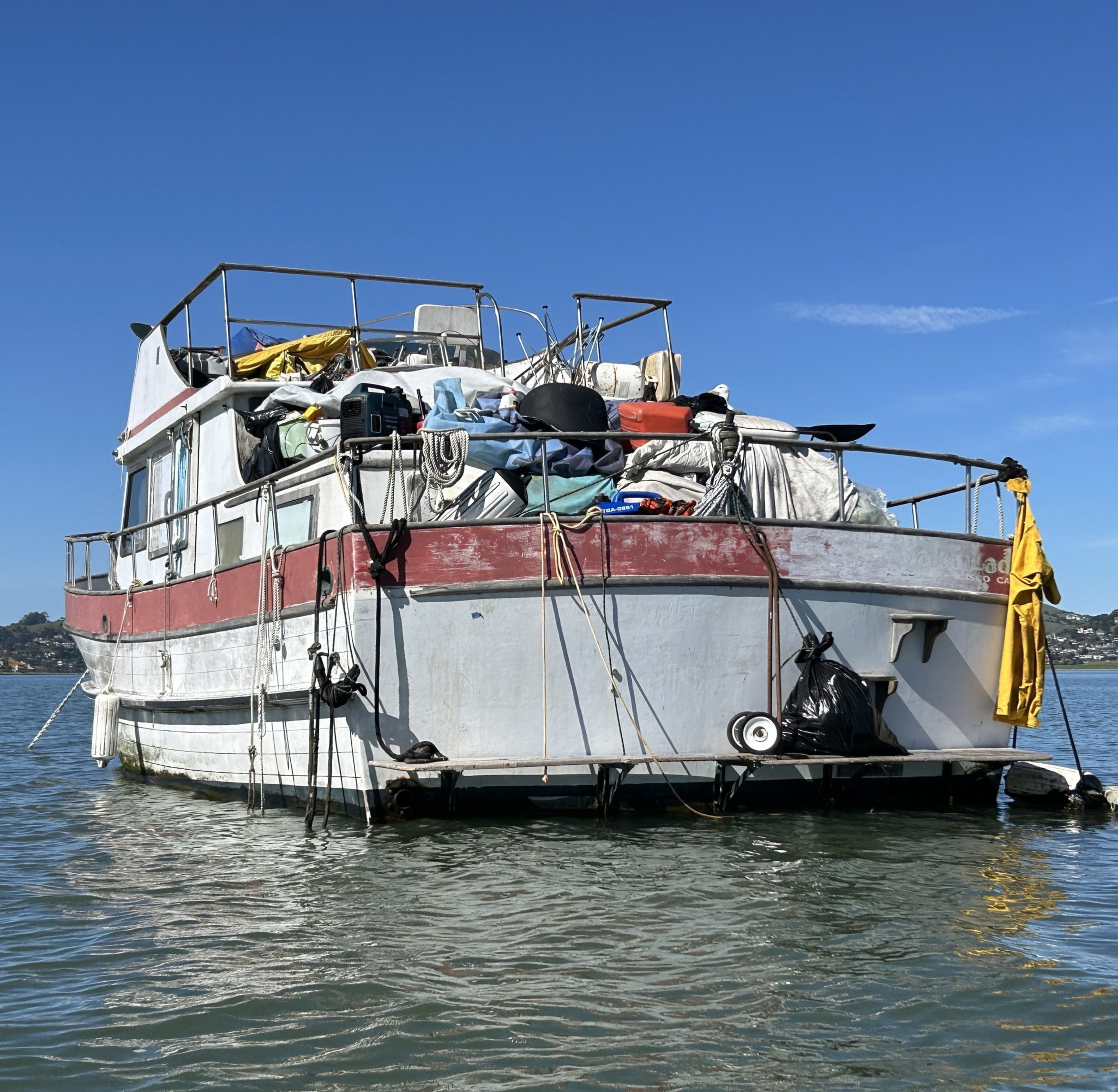 An old boat heavily cluttered with items, floating on calm water under a clear blue sky.