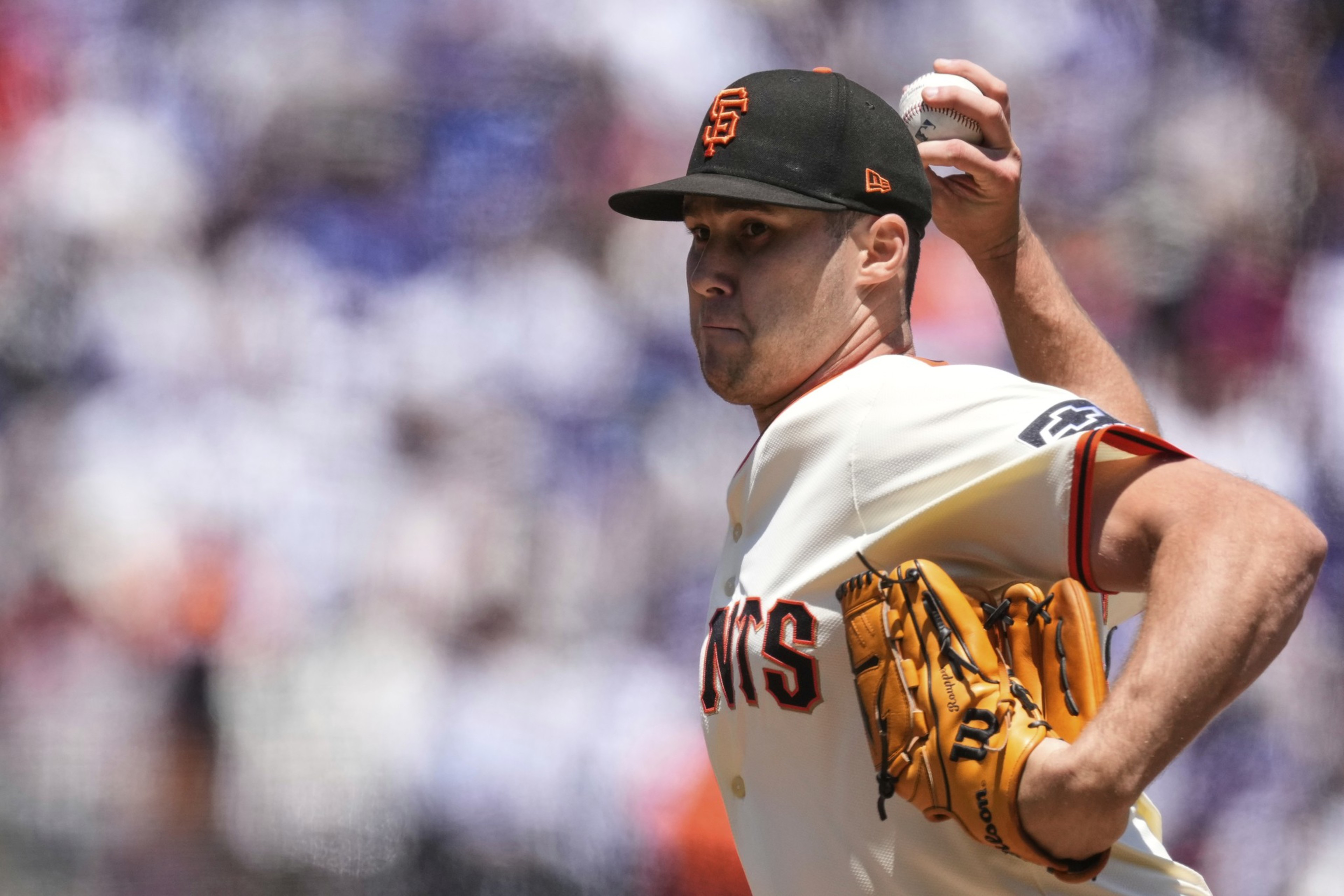 A baseball player in a Giants uniform is pitching. He's wearing an orange glove and a black cap with an &quot;SF&quot; logo, against a blurred crowd backdrop.