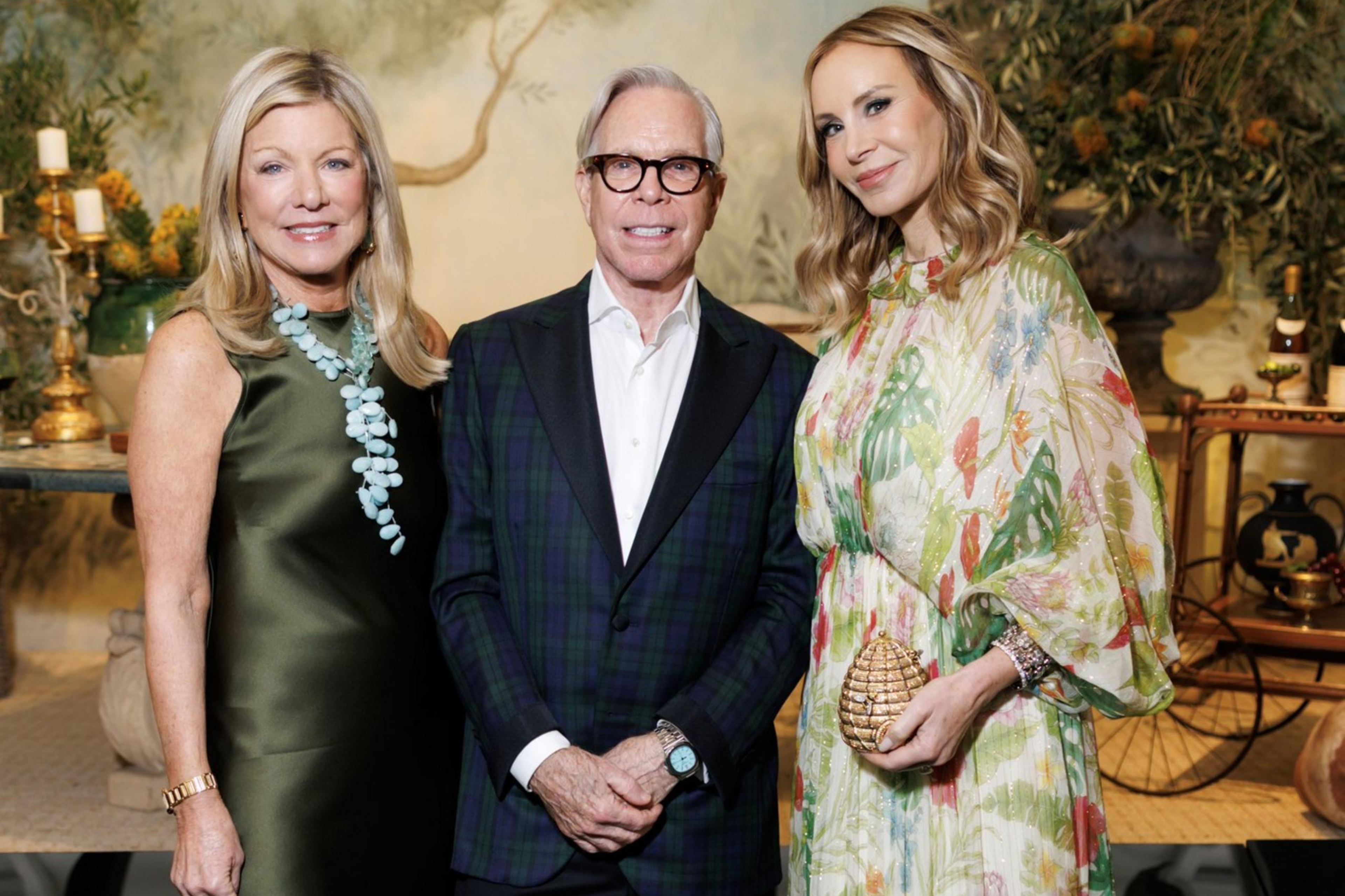 A man in a dark plaid blazer stands between two women; one in a green satin dress with a turquoise necklace, the other in a floral, flowing dress holding a gold clutch.