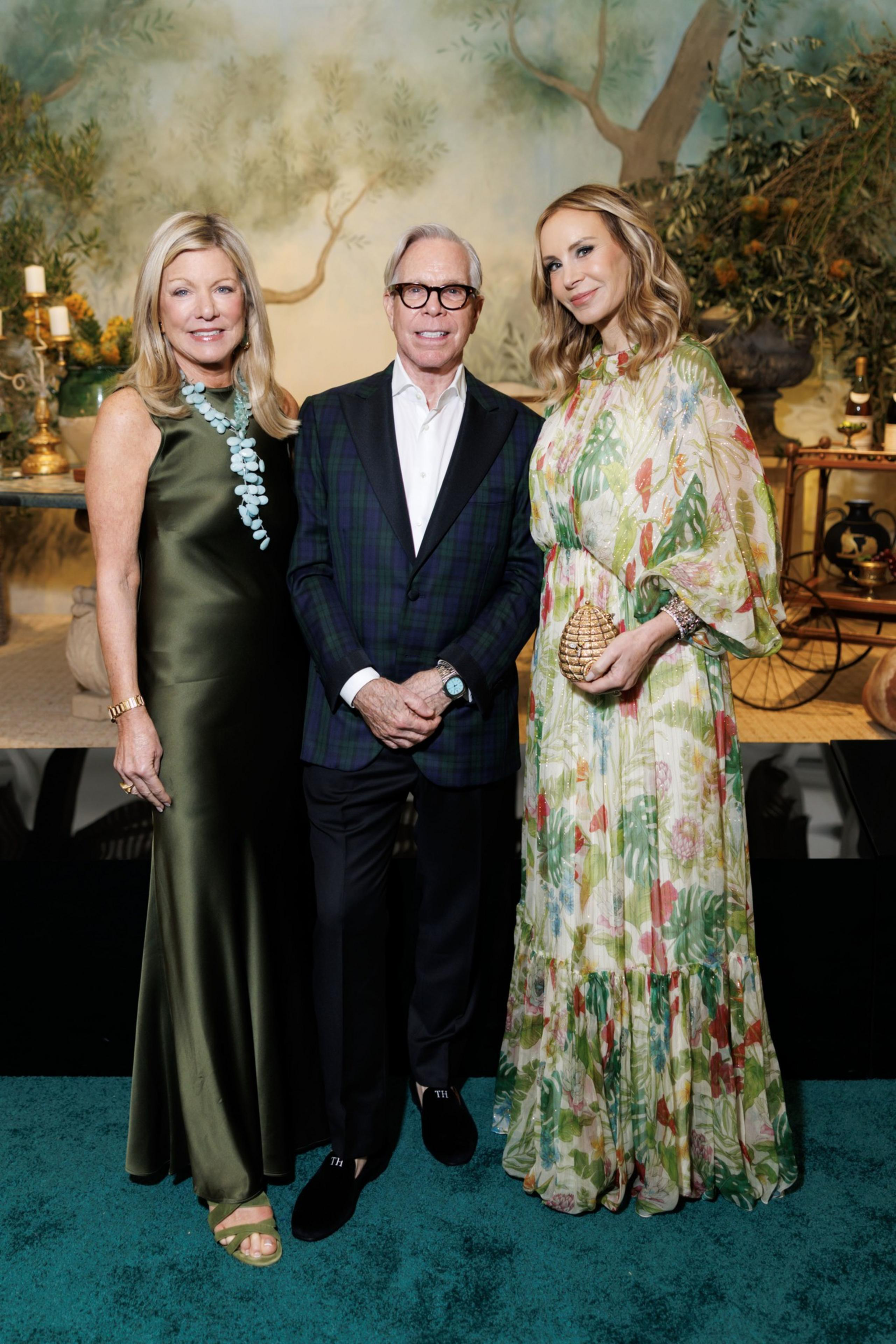 A man in a dark plaid blazer stands between two women; one in a green satin dress with a turquoise necklace, the other in a floral, flowing dress holding a gold clutch.