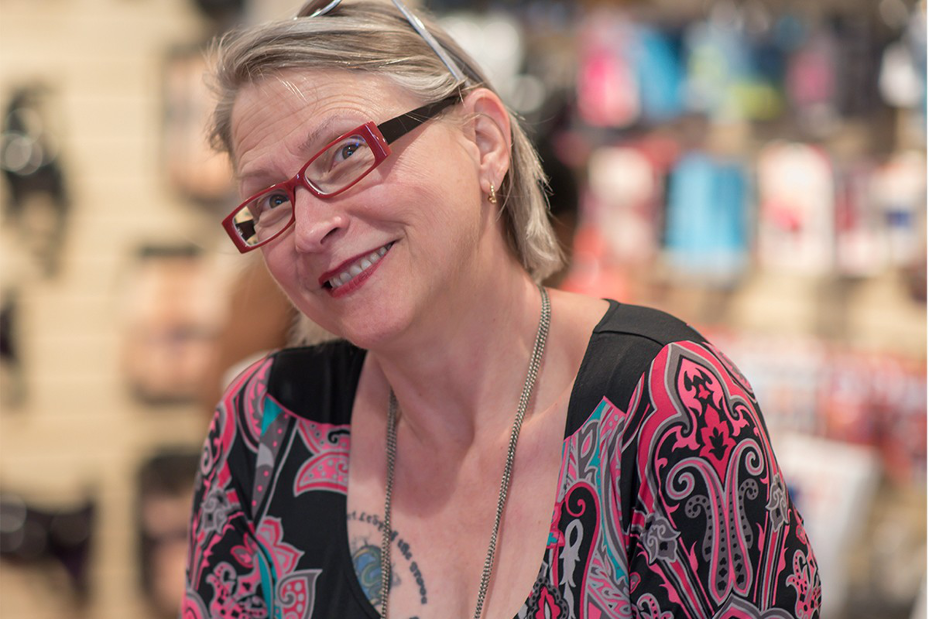 A woman with short hair and red glasses smiles at the camera. She wears a colorful patterned top and a necklace, with blurred shelves in the background.